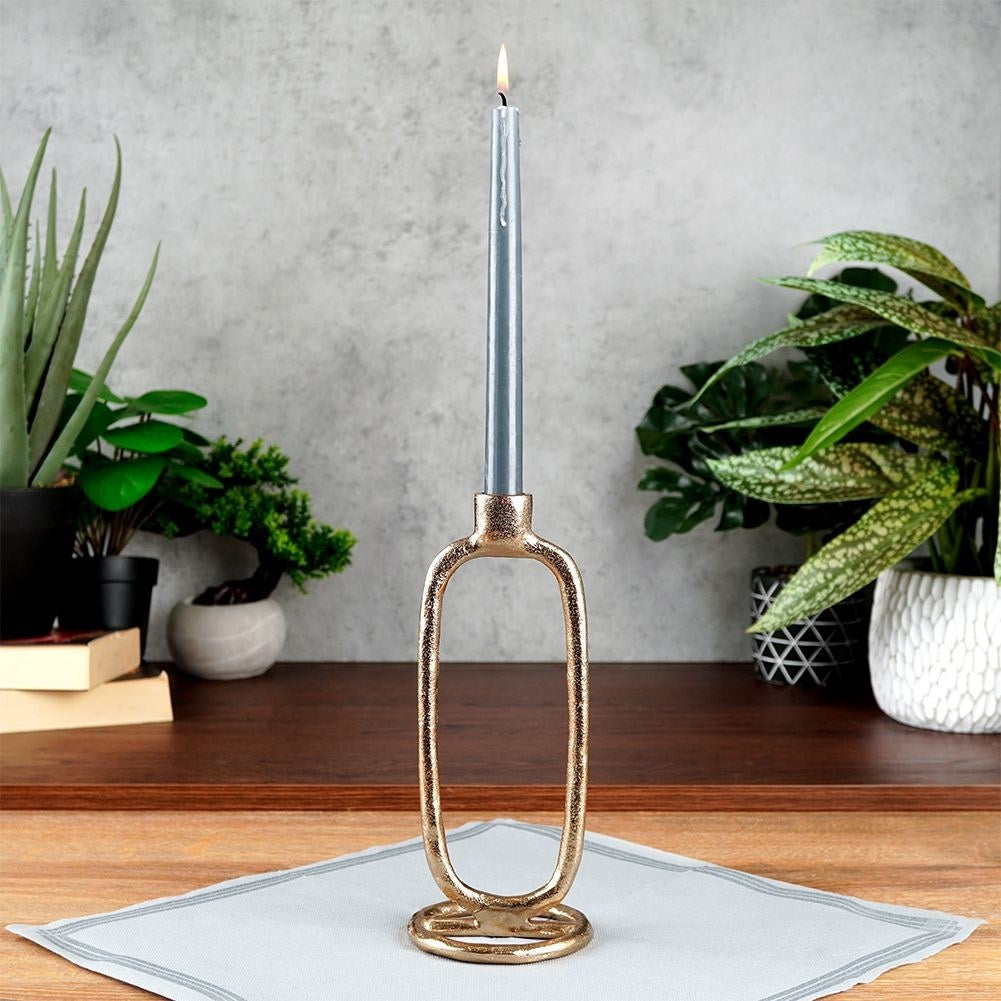 Una vela gris encendida en un soporte dorado se encuentra sobre una servilleta gris claro. Hay plantas en el fondo. La escena es interior.