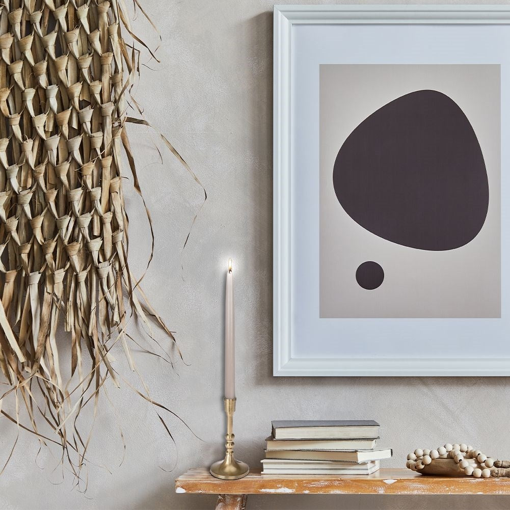 Una vela encendida en un soporte de latón, obras de arte con formas oscuras, decoración de pared tejida, libros y un cuenco de madera con cuentas en un estante.