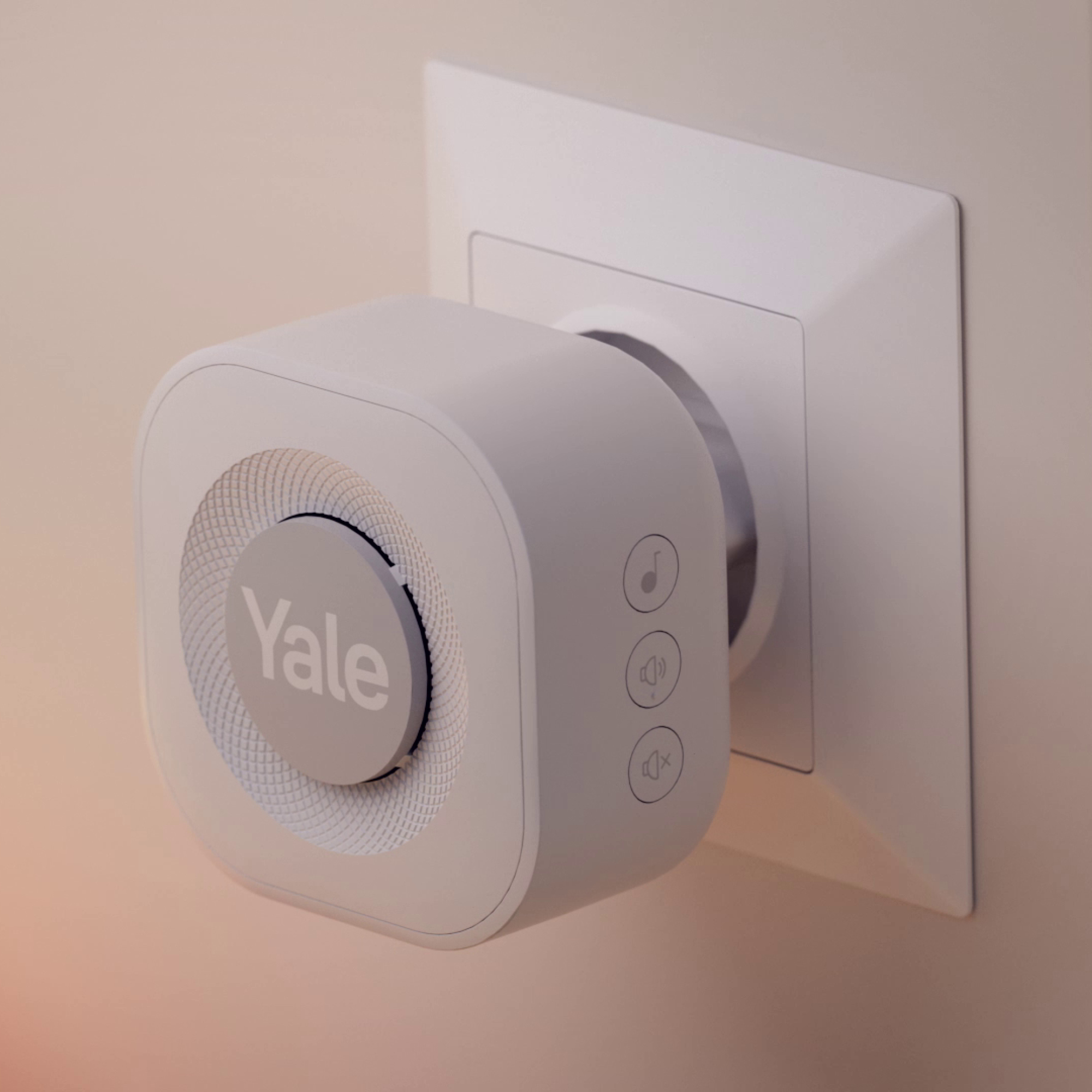 Una spina intelligente Yale bianca in una presa a muro. Ha un altoparlante e pulsanti di controllo. Il muro è beige chiaro.