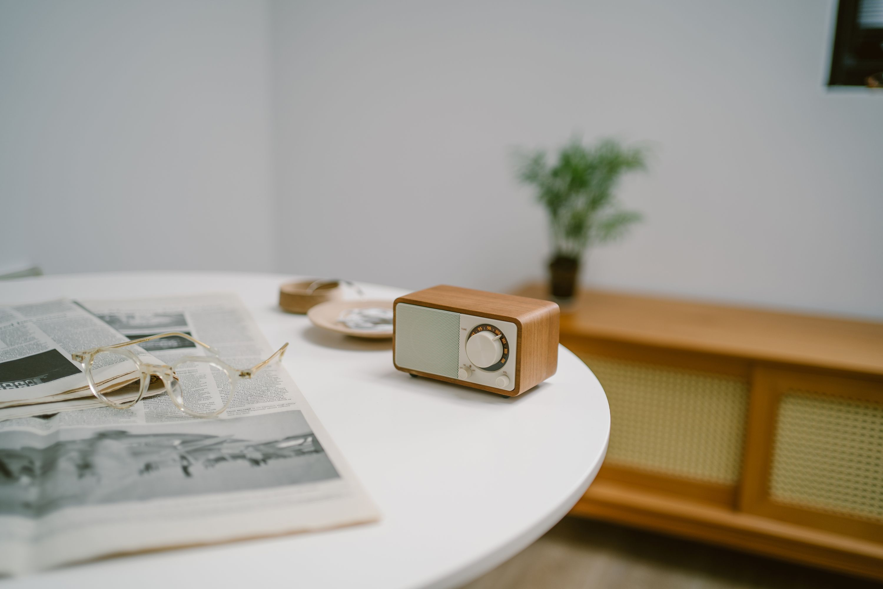 Ein Holzradio steht auf einem weißen Tisch mit Zeitung und Brille. Ein Blumentopf und Schrank sind im Hintergrund.
