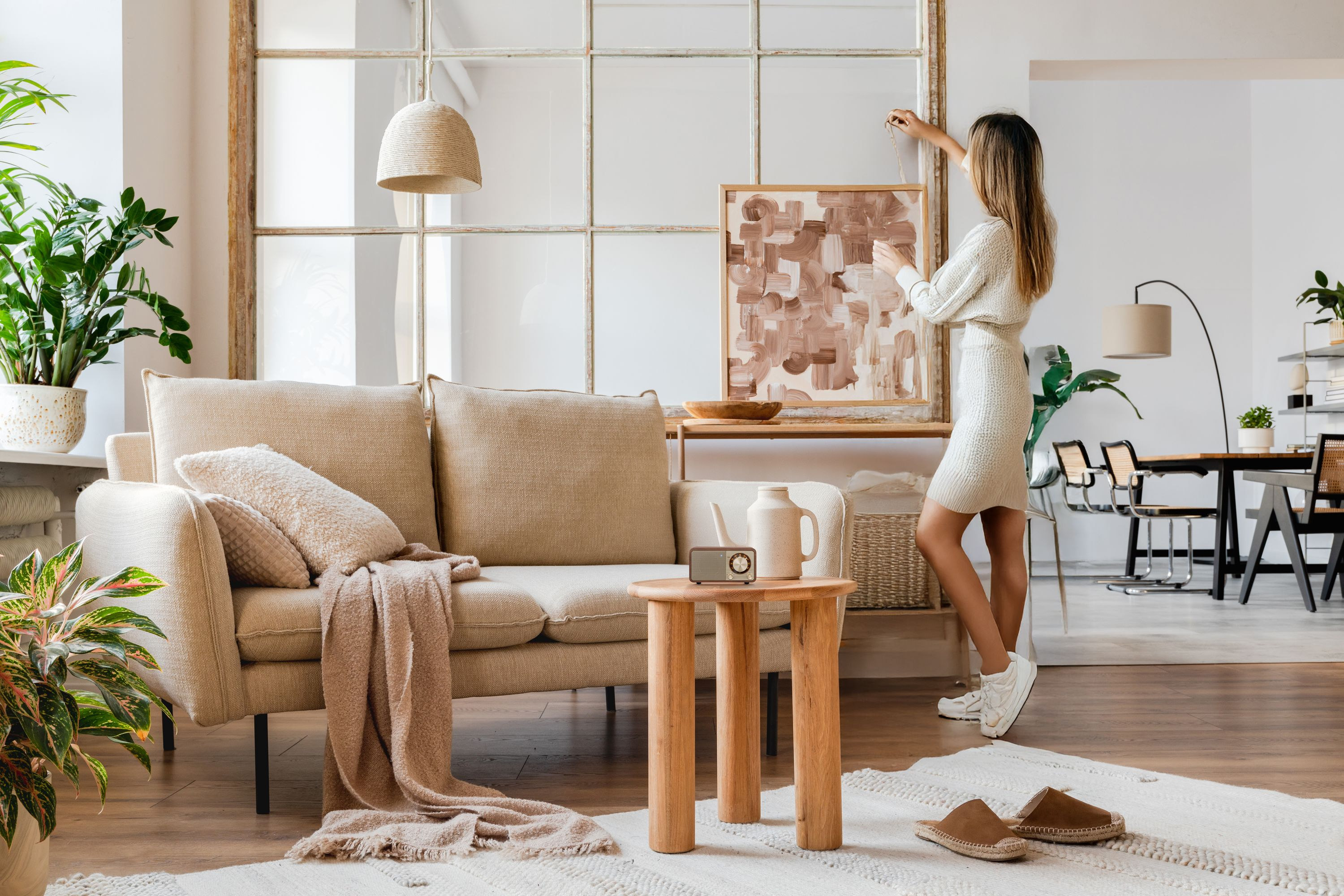 Eine Frau richtet ein Gemälde in einem Wohnzimmer aus. Sofa, Tisch, Pflanzen und Teppich sind sichtbar. Helle Farben dominieren.