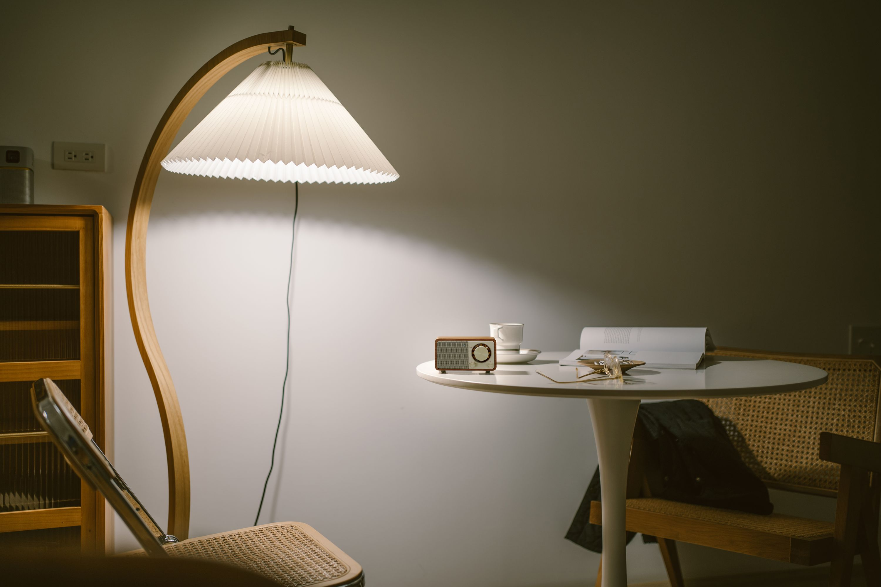 Ein Zimmer mit Lampe, Tisch, Radio, Stuhl und Schrank. Die Lampe ist an. Eine Tasse, ein Buch und eine Brille auf dem Tisch.