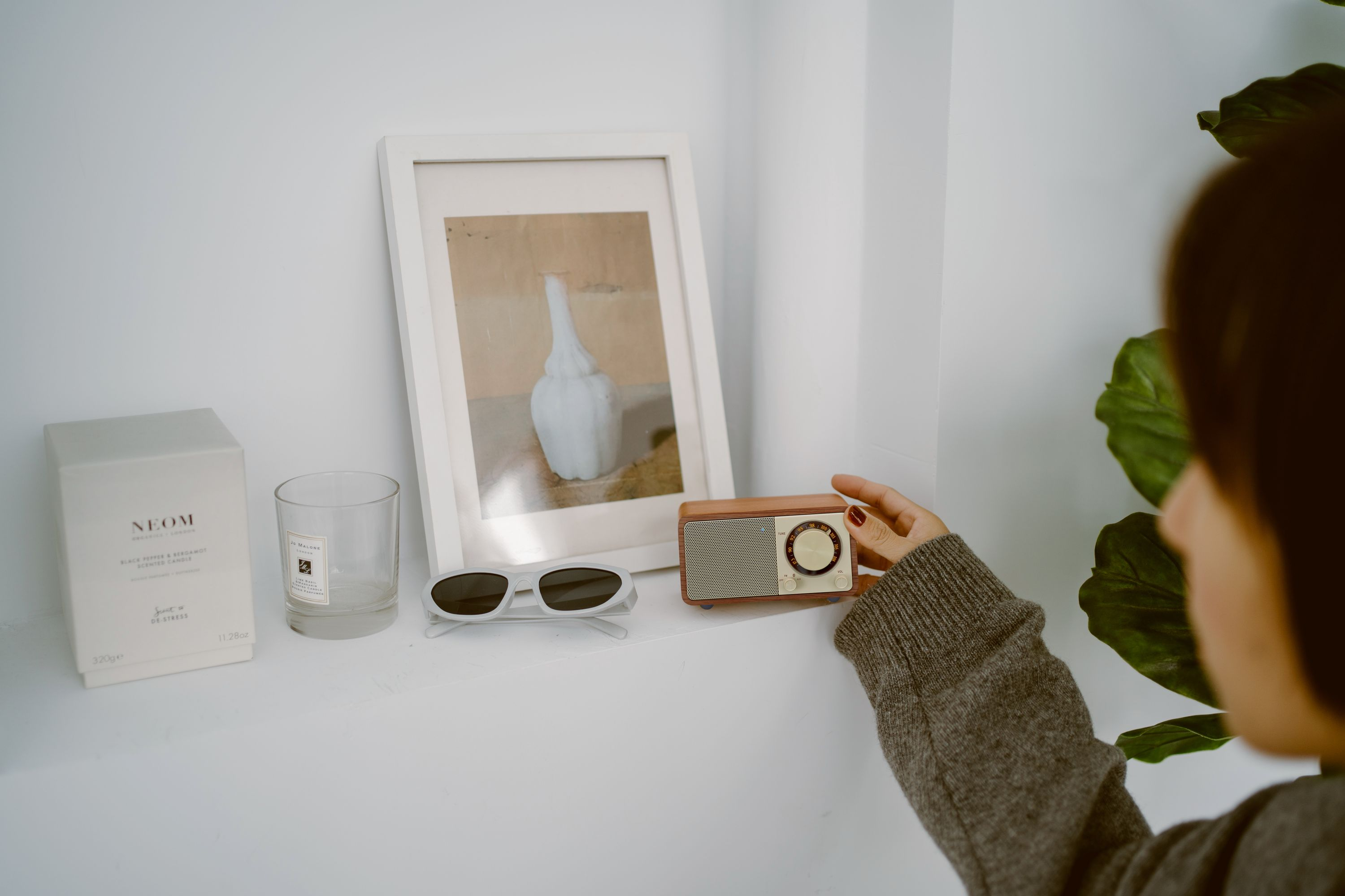 Eine Hand berührt ein Holzradio auf einem weißen Regal. Sonnenbrille, Kerze und ein gerahmtes Bild sind in der Nähe.