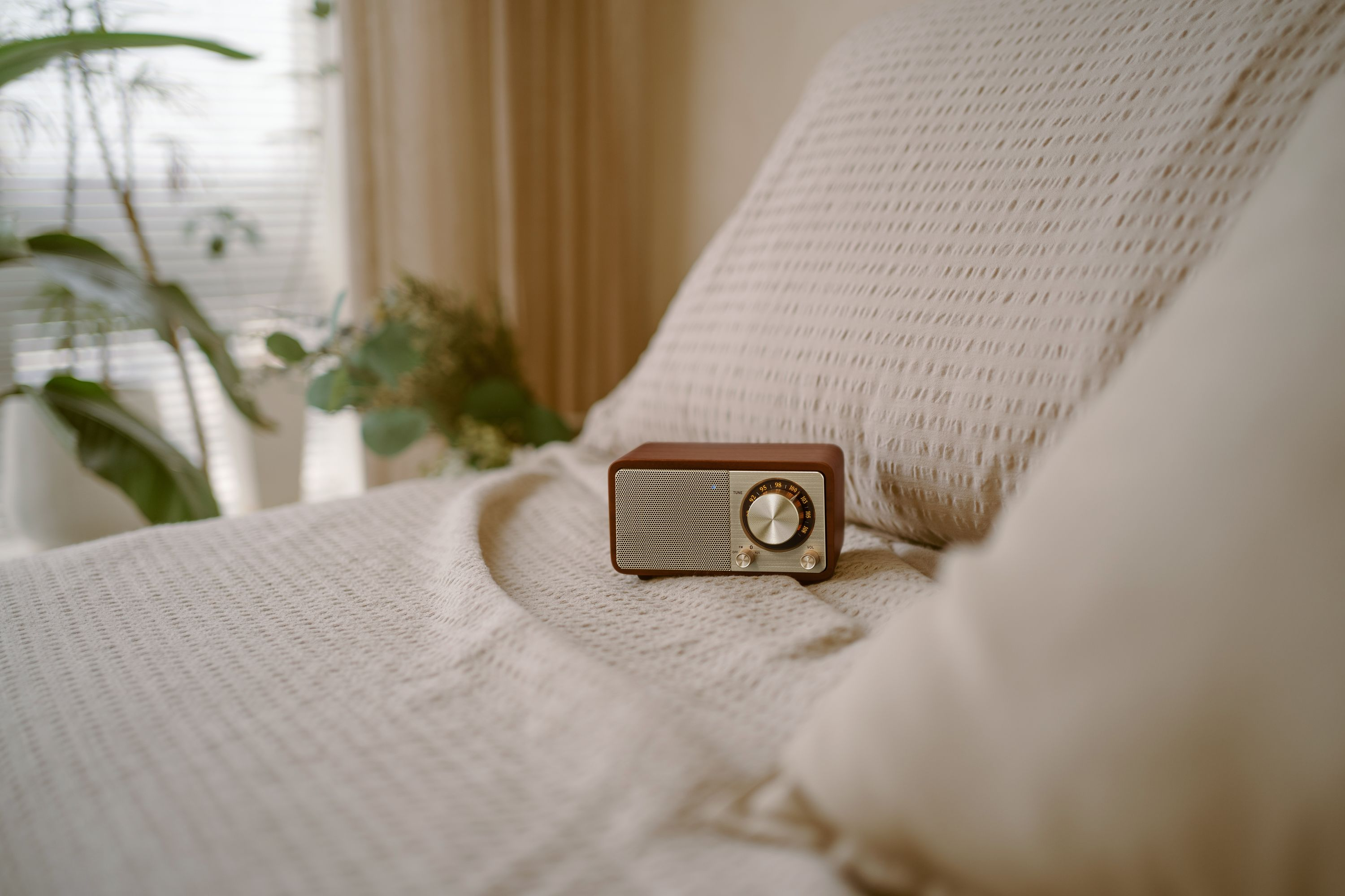 Ein braunes Radio auf einem beigefarbenen Bett. Im Hintergrund ein Fenster mit Jalousien und Pflanzen. Beige Vorhänge sind ebenfalls zu sehen.