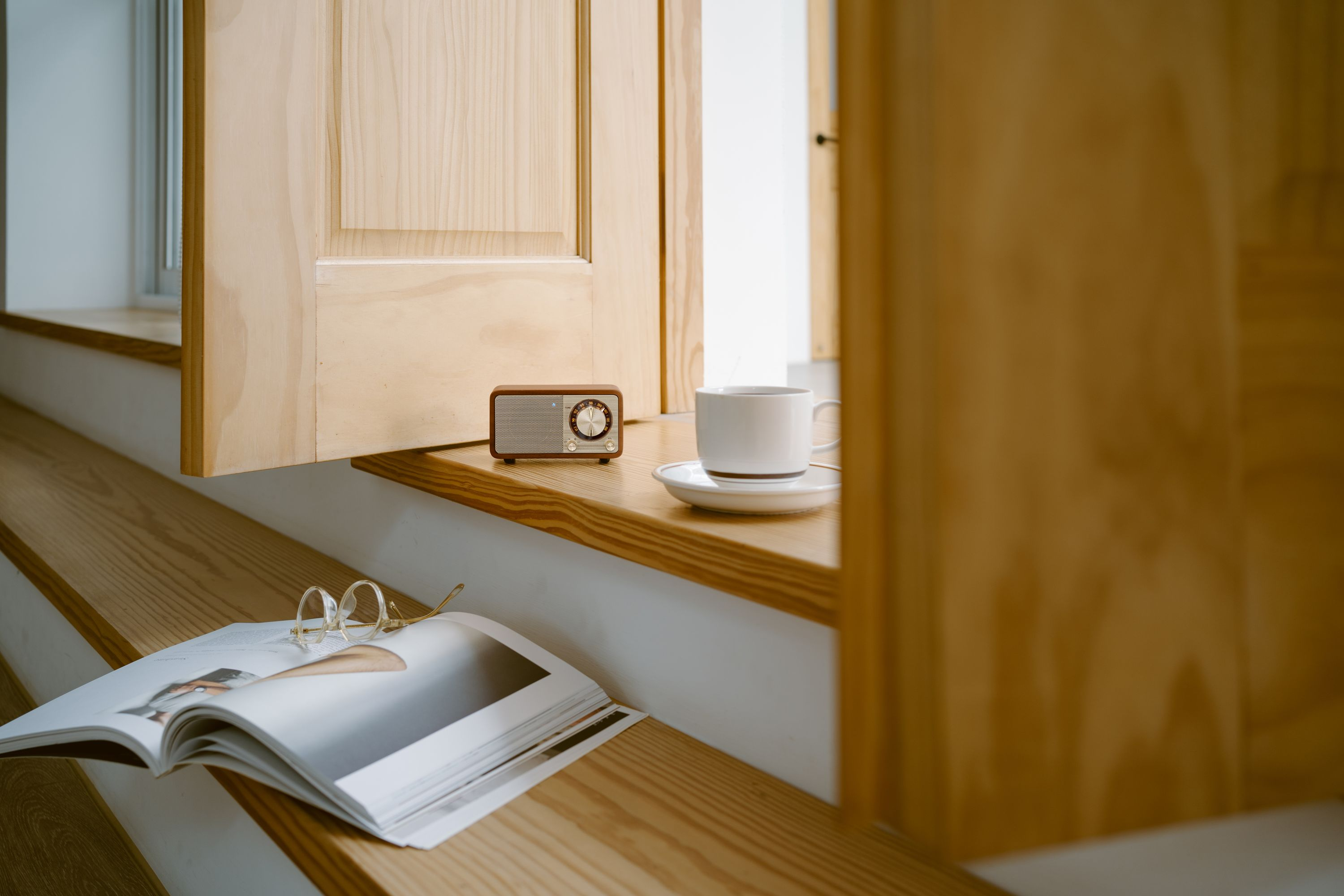 Ein Holzradio, eine Kaffeetasse, ein Buch und eine Brille auf einer Holzoberfläche in der Nähe eines Fensters.