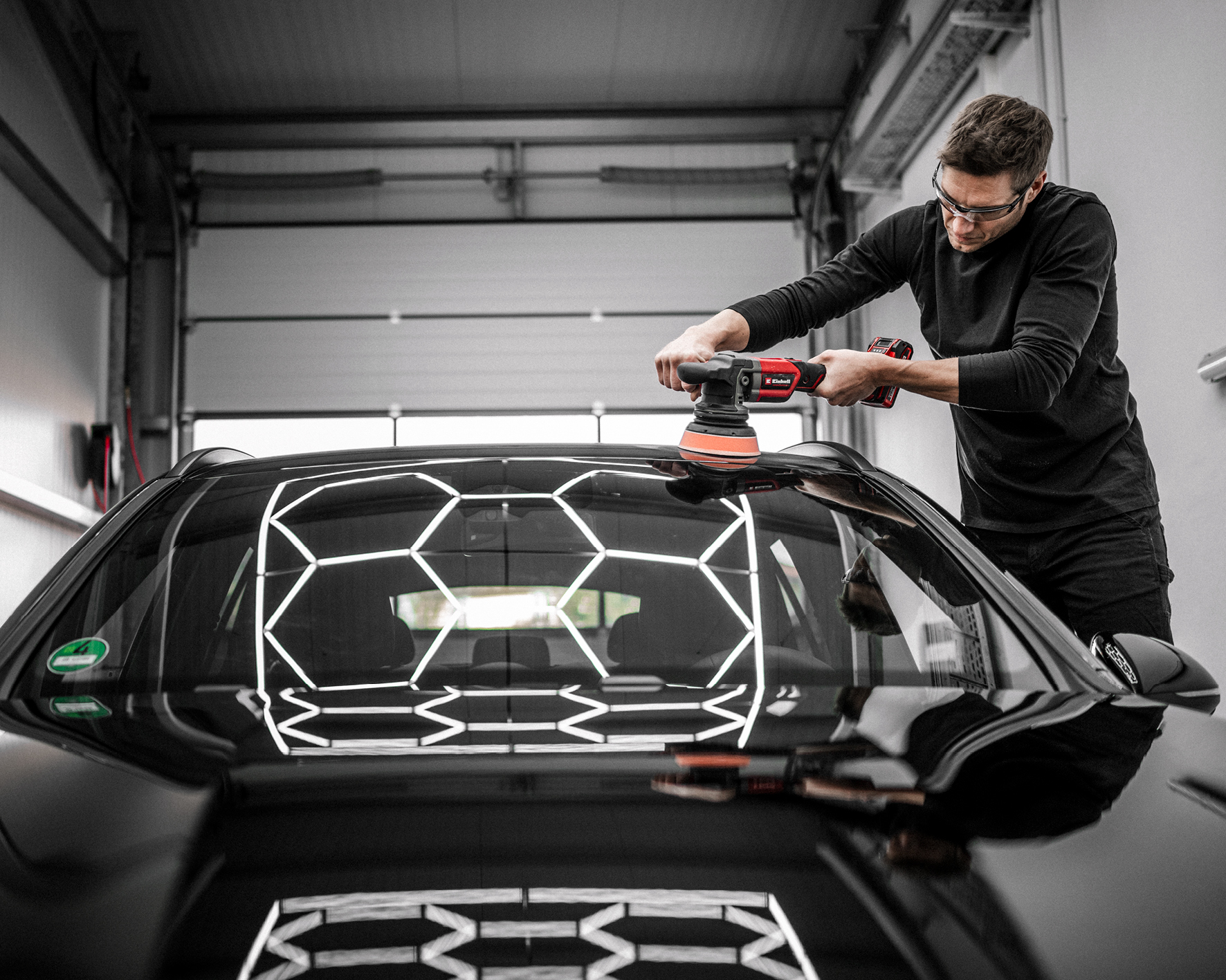 Ein Mann poliert ein schwarzes Auto mit einer Maschine. Das Auto steht in einer Garage. Auf der Oberfläche des Autos sind Reflexlichter zu sehen.