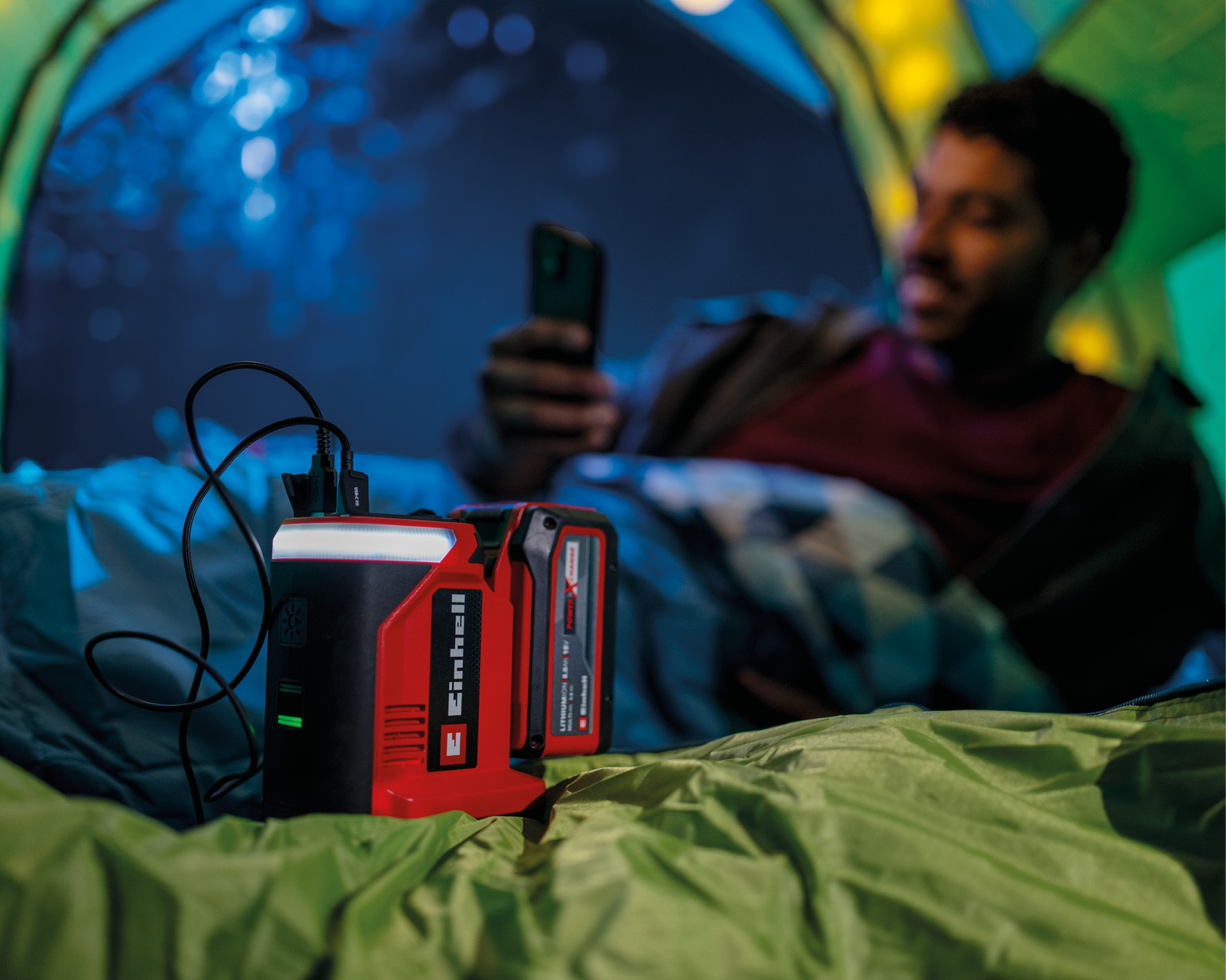 Ein Mann im Zelt benutzt sein Handy, im Vordergrund eine rot-schwarze Powerbank. Es ist mit einem Kabel verbunden.