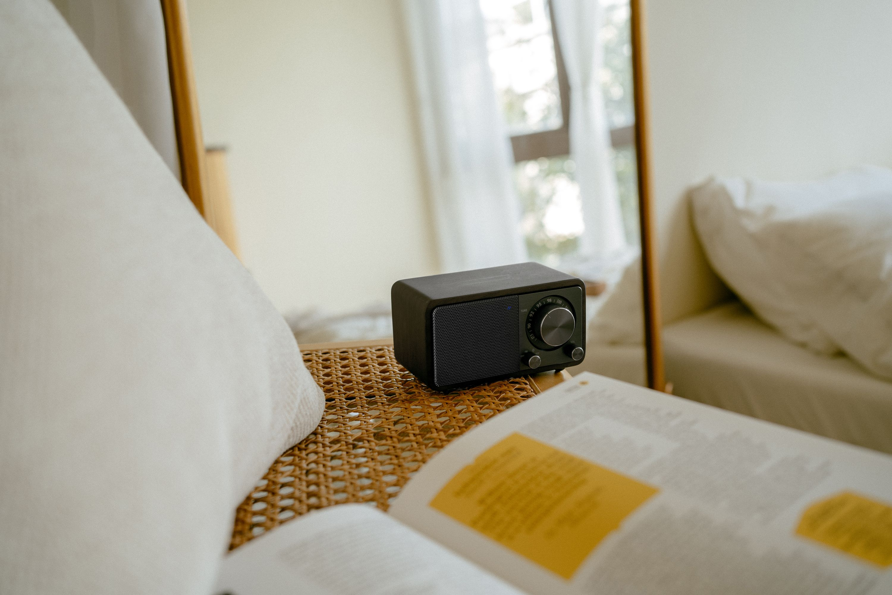 Ein schwarzes Radio steht auf einem Korbtisch in der Nähe eines offenen Buches. Ein Spiegel reflektiert ein Fenster und ein Bett.