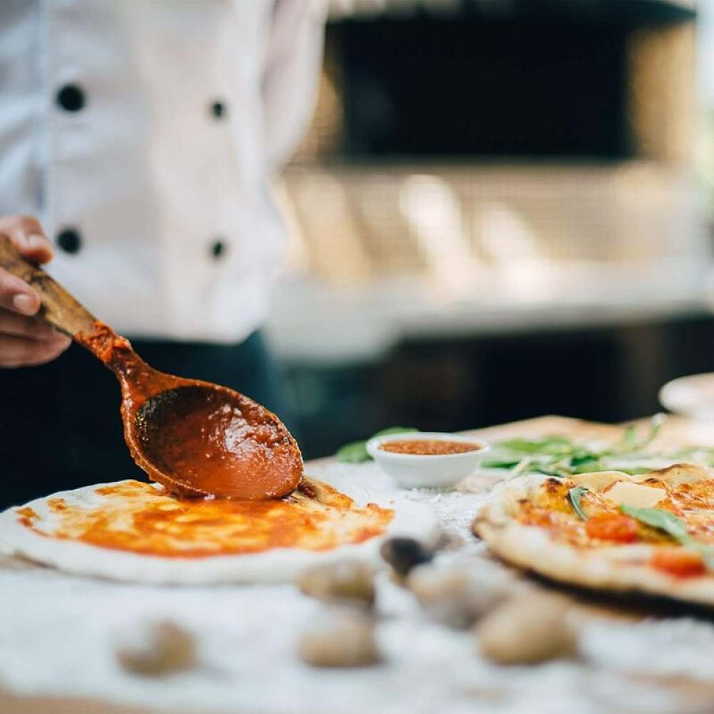 Ein Koch verteilt Tomatensauce mit einem Holzlöffel auf Pizzateig. Eine weitere Pizza liegt im Hintergrund, bereit zum Backen.