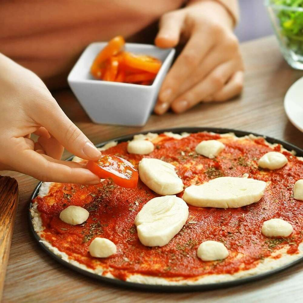 Eine Person legt eine Tomatenscheibe auf eine Pizza mit Mozzarella. Es gibt eine Schüssel mit Tomaten und Kräutern auf der Pizza. Der Tisch ist aus Holz.
