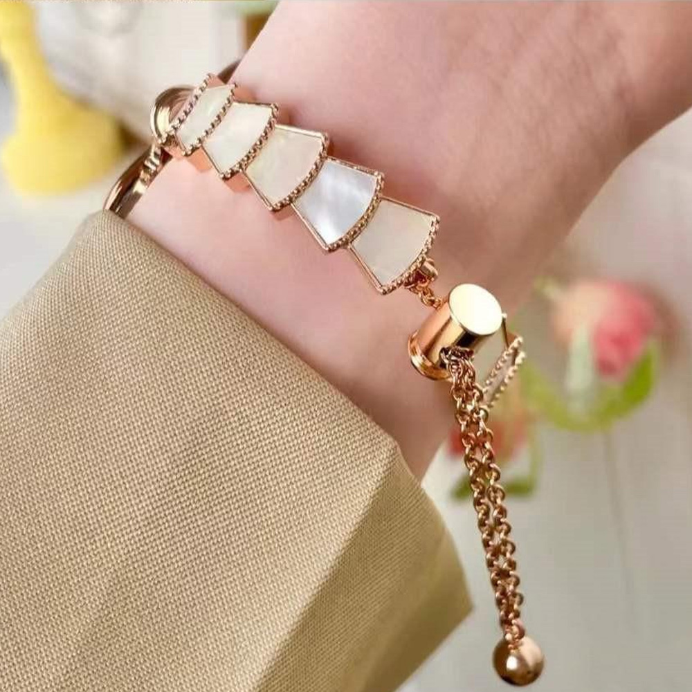 Eine Hand trägt ein goldenes Armband mit weiß muschelartigen Formen. Das Armband befindet sich an einem Handgelenk, mit einem beigen Ärmel und einem verschwommenen Hintergrund.