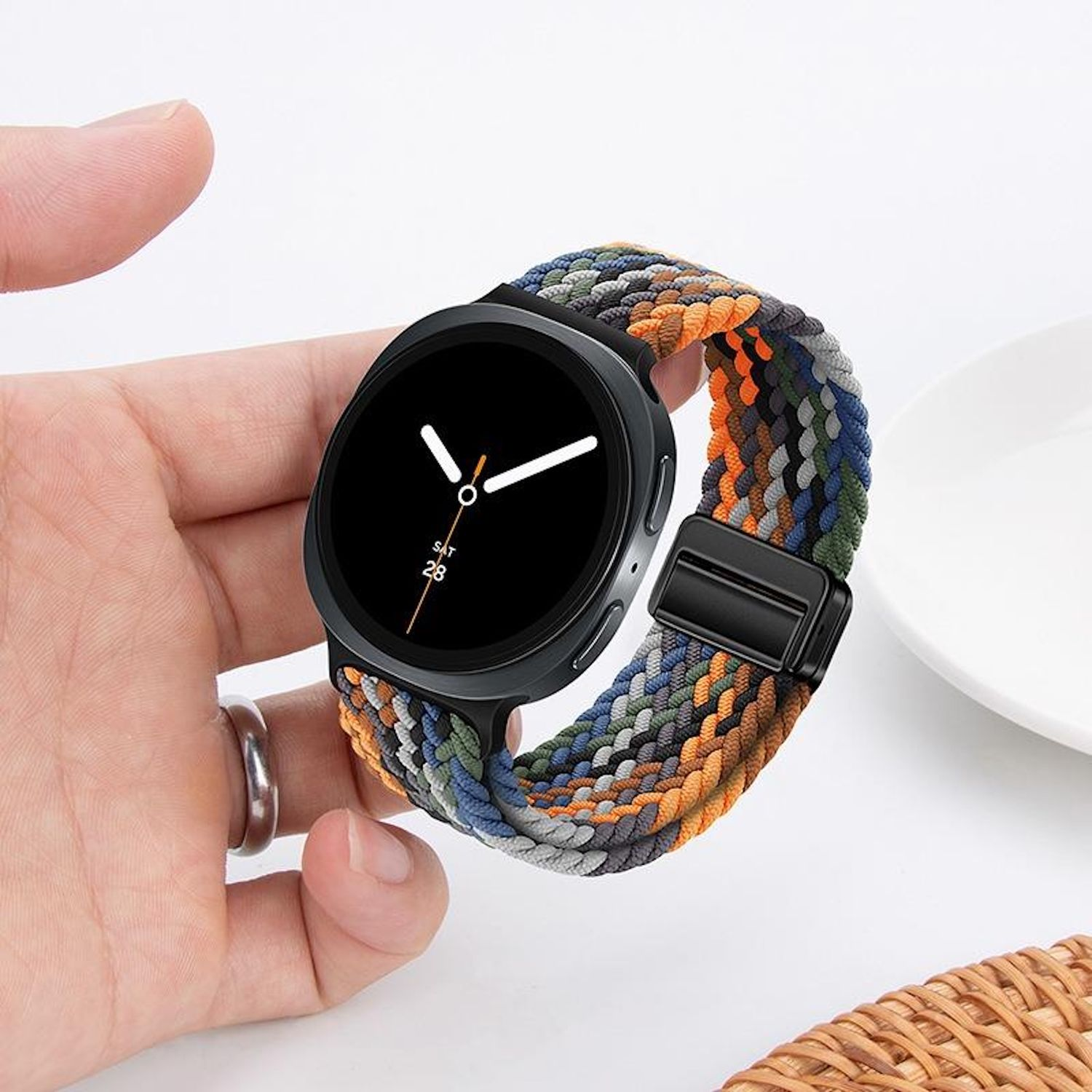 Eine schwarze Smartwatch mit einem bunten geflochtenen Armband wird von einer Hand mit einem silbernen Ring gehalten. Ein weißer Teller und ein Korb sind im Hintergrund.