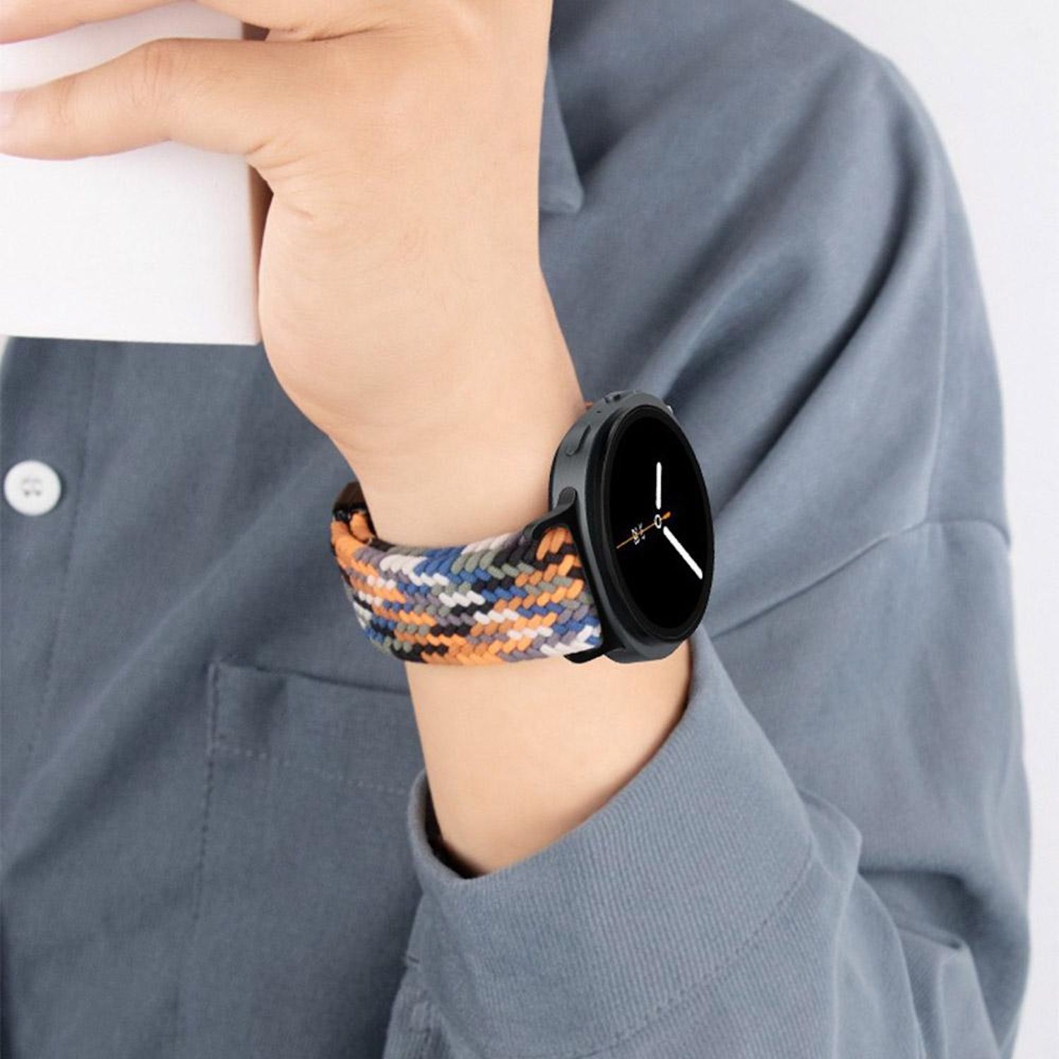 Eine Person trägt eine Smartwatch mit einem bunten geflochtenen Armband. Die Uhr ist schwarz. Die Person trägt ein graues Hemd.