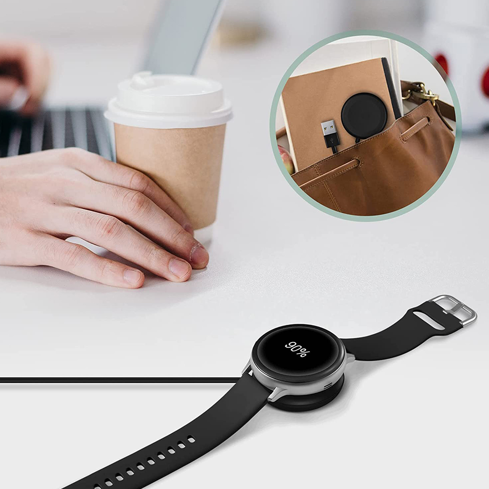 Eine Smartwatch wird auf einer weißen Oberfläche aufgeladen. Eine Kaffeetasse und eine Tasche mit Ladegerät sind ebenfalls sichtbar.