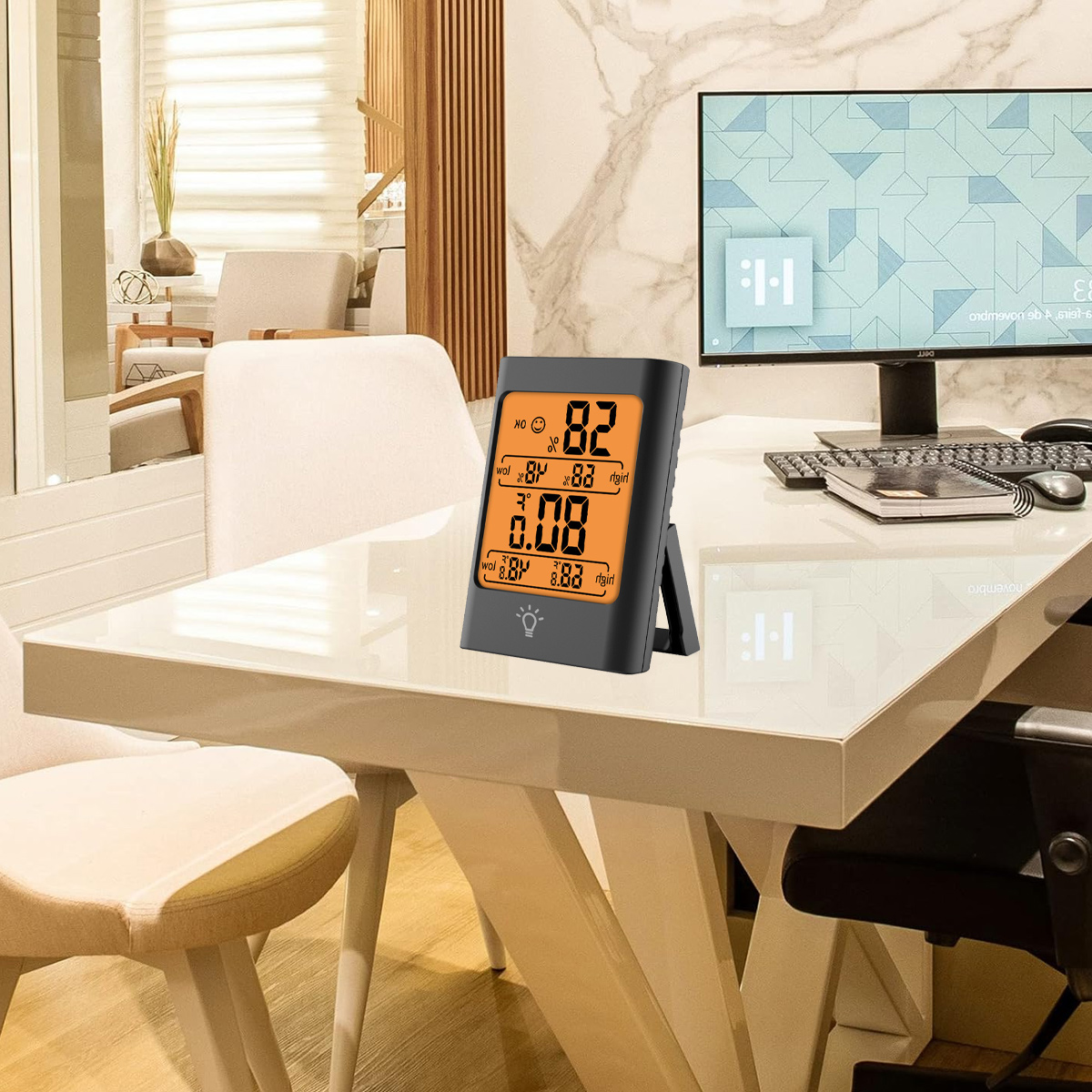 Ein digitales Thermometer zeigt Temperatur und Luftfeuchtigkeit auf einem weißen Schreibtisch an. Ein Computerbildschirm und Büromöbel sind im Hintergrund zu sehen.