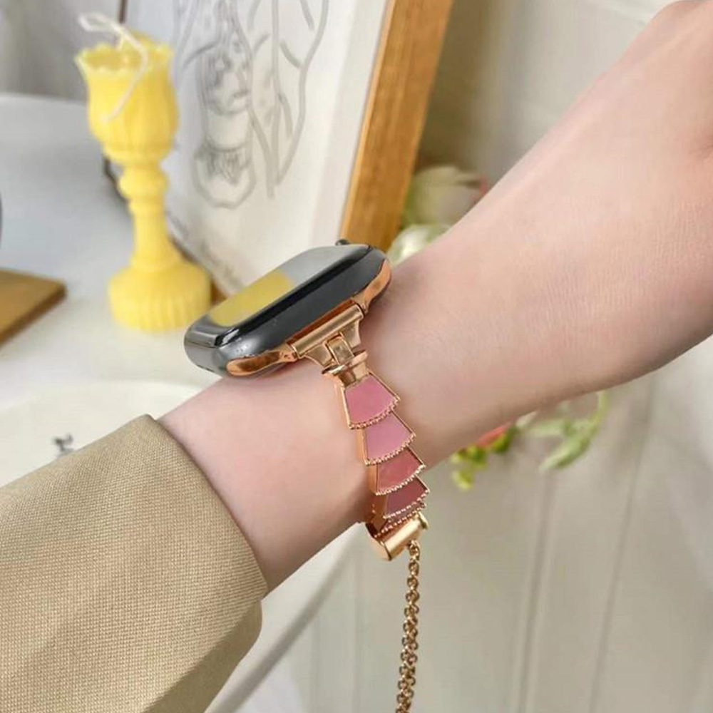 Handgelenk mit roségoldener Smartwatch und pinkfarbenem, gestuftem Armband. Gelbe Kerze und Bilderrahmen im Hintergrund.