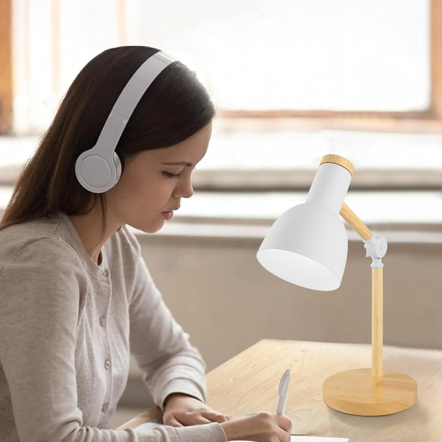 Frau mit Kopfhörern schreibt. Eine weiße Schreibtischlampe steht auf dem Tisch. Die Lampe hat einen Holzfuß und -arm.