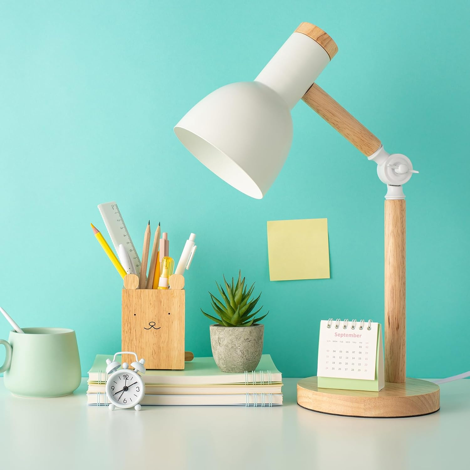 Ein Schreibtisch mit weißer Lampe, Schreibwaren, einer kleinen Uhr, Kalender, Becher und einer Pflanze vor türkisem Hintergrund.