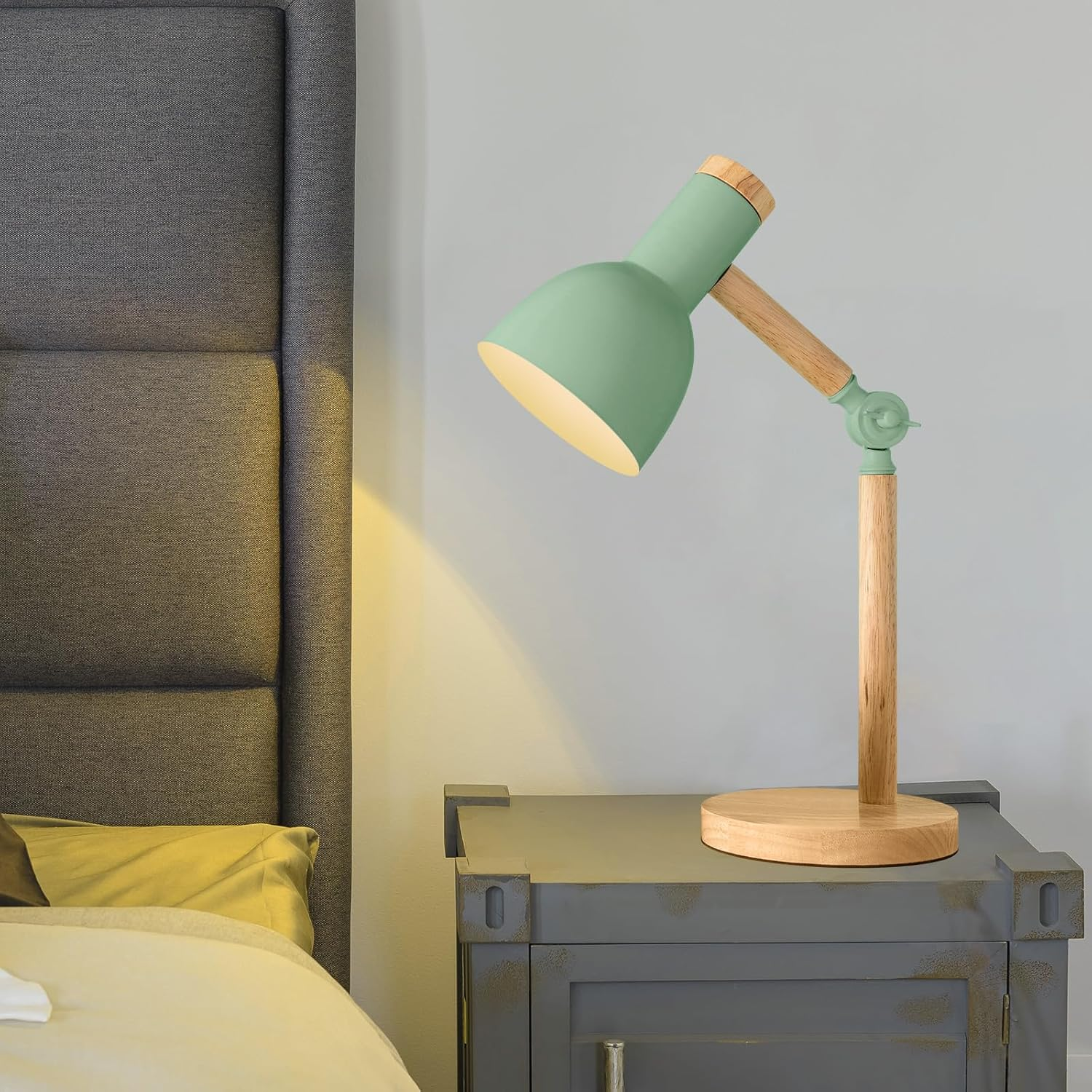Eine grüne Schreibtischlampe mit Holzakzenten steht auf einem grauen Nachttisch. Im Hintergrund ist ein Bett mit grauem Kopfteil.
