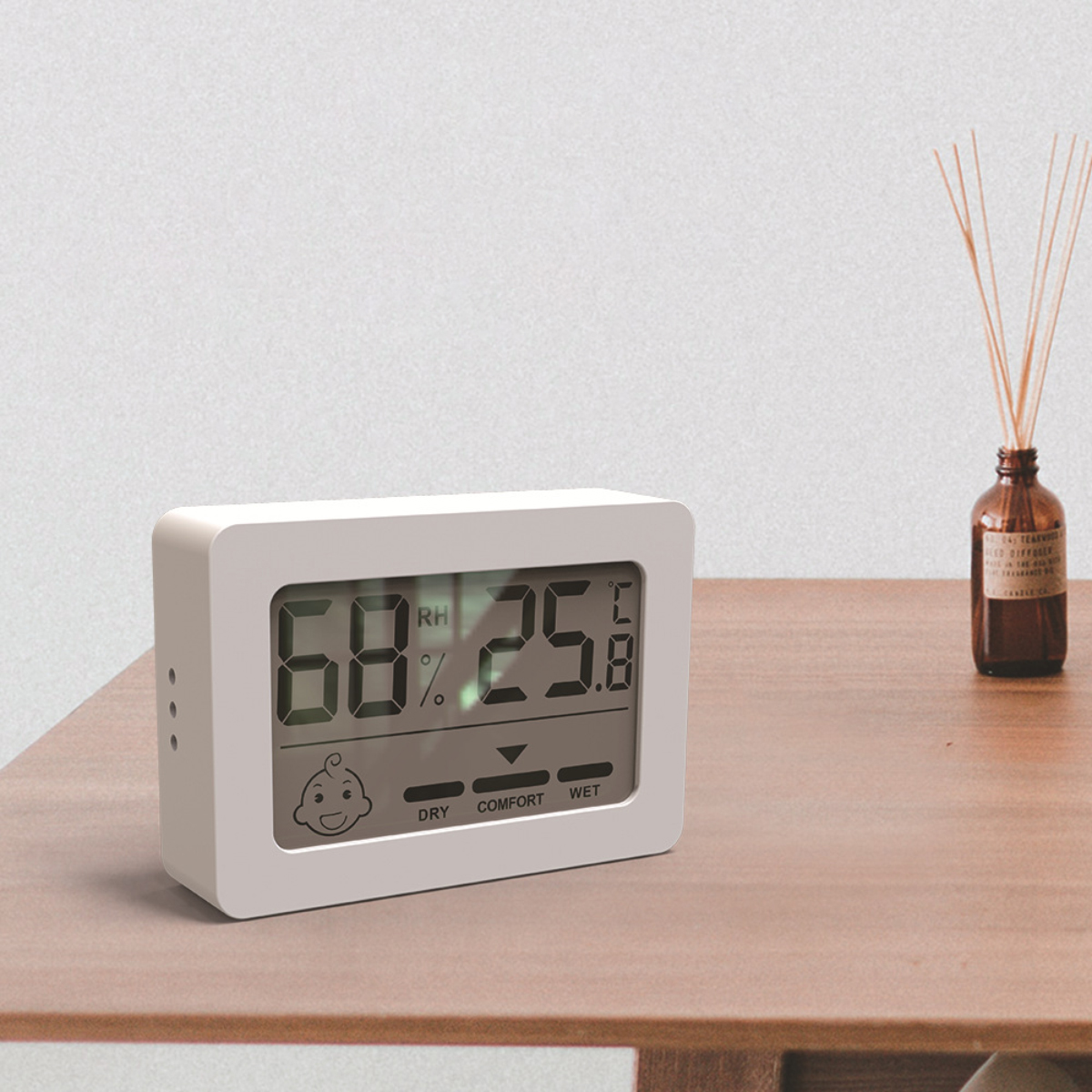 Ein weißes digitales Thermometer zeigt Luftfeuchtigkeit und Temperatur an. Ein Baby-Symbol ist abgebildet. Rechts steht eine braune Flasche mit Stäbchen.