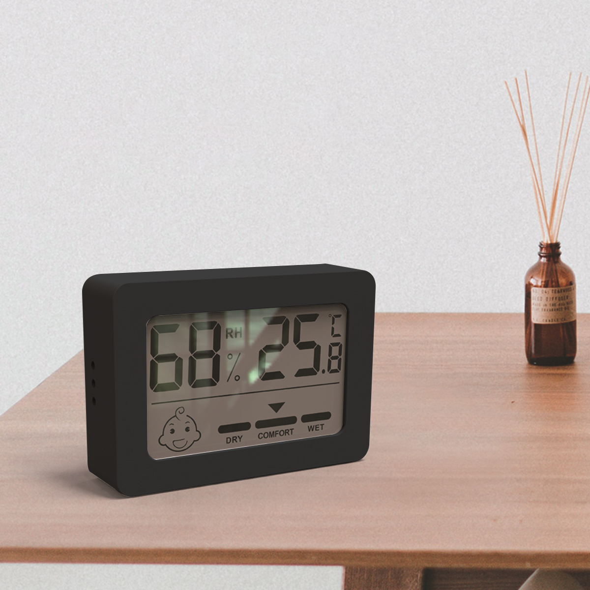 Ein schwarzes digitales Hygrometer zeigt 68 % Luftfeuchtigkeit und 25,8 °C an. Es steht auf einem Holztisch neben einer braunen Glasflasche mit Schilf.
