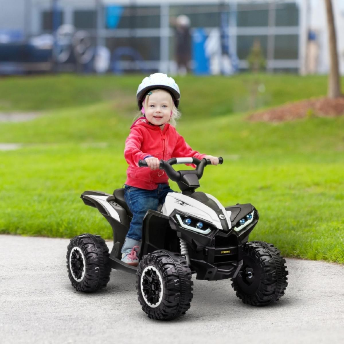 Ein lächelndes Kleinkind in roter Jacke und Helm fährt auf einem schwarzen und weißen Quad auf einem gepflasterten Weg, mit grünem Gras im Hintergrund.