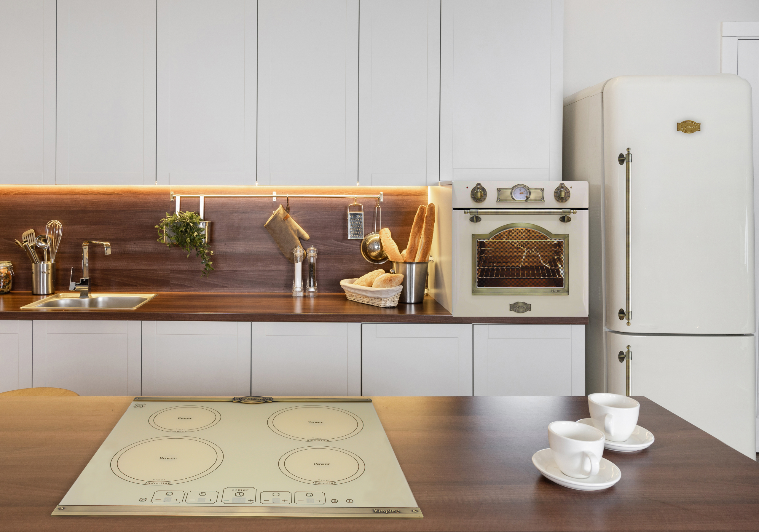 Eine Küchenszene mit Herd, Backofen, Kühlschrank und zwei Tassen. Weiße Schränke und braune Holz-Arbeitsplatte.