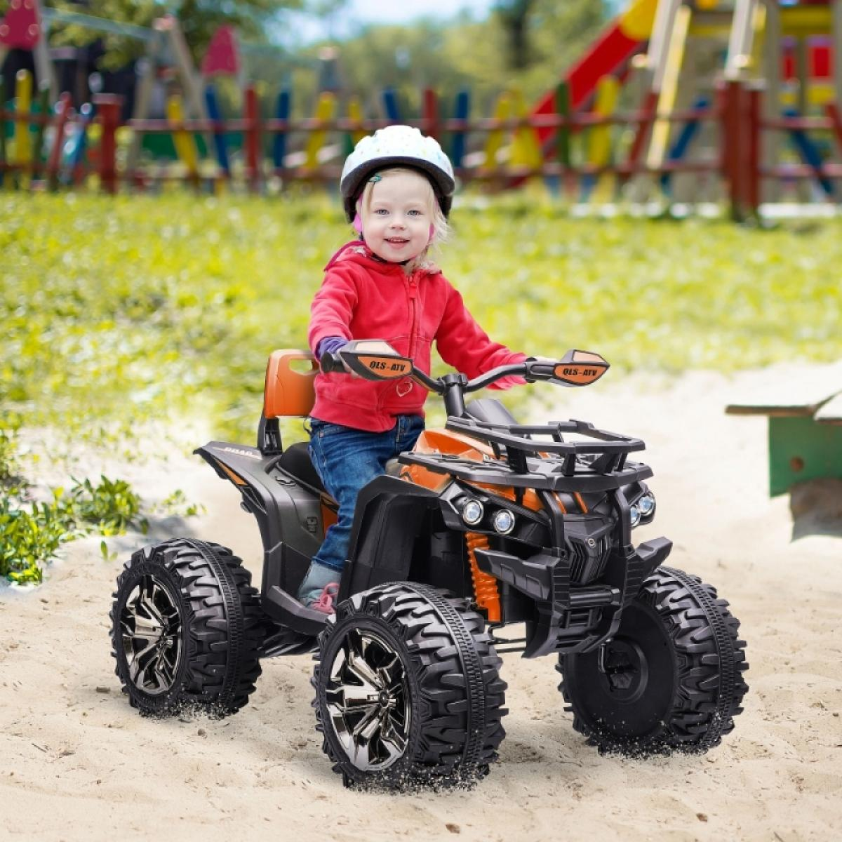 Ein Kind in roter Jacke und Helm fährt auf Sand ein orange-schwarzes ATV. Im Hintergrund sind Spielplatz und Grün.