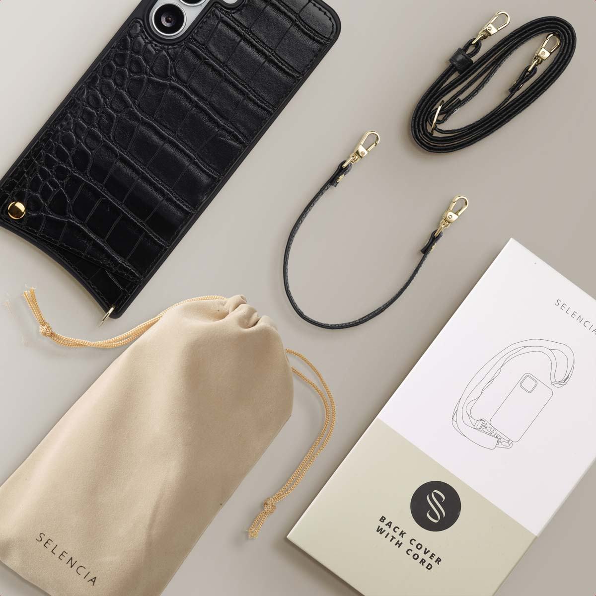 Zwarte telefoonhoes met riem, een beige zakje met 'Selencia' en een witte doos 'Back cover with cord' op een grijs oppervlak.