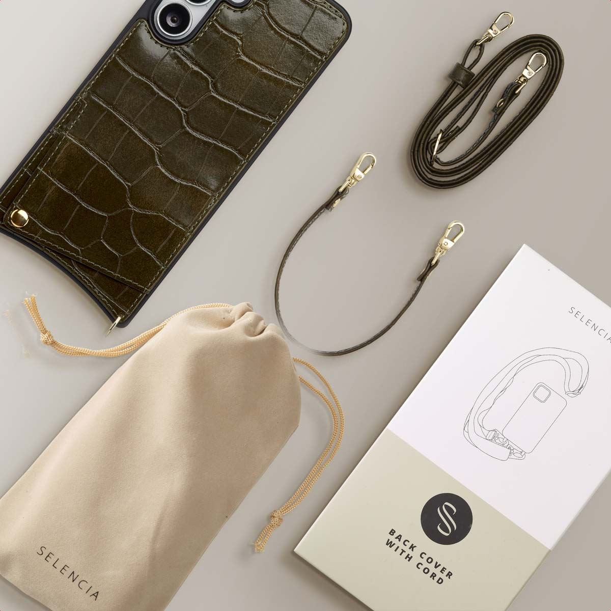 Olijfgroene telefoonhoes, koord, zakje en verpakking op grijs oppervlak. Items zijn diagonaal gerangschikt.