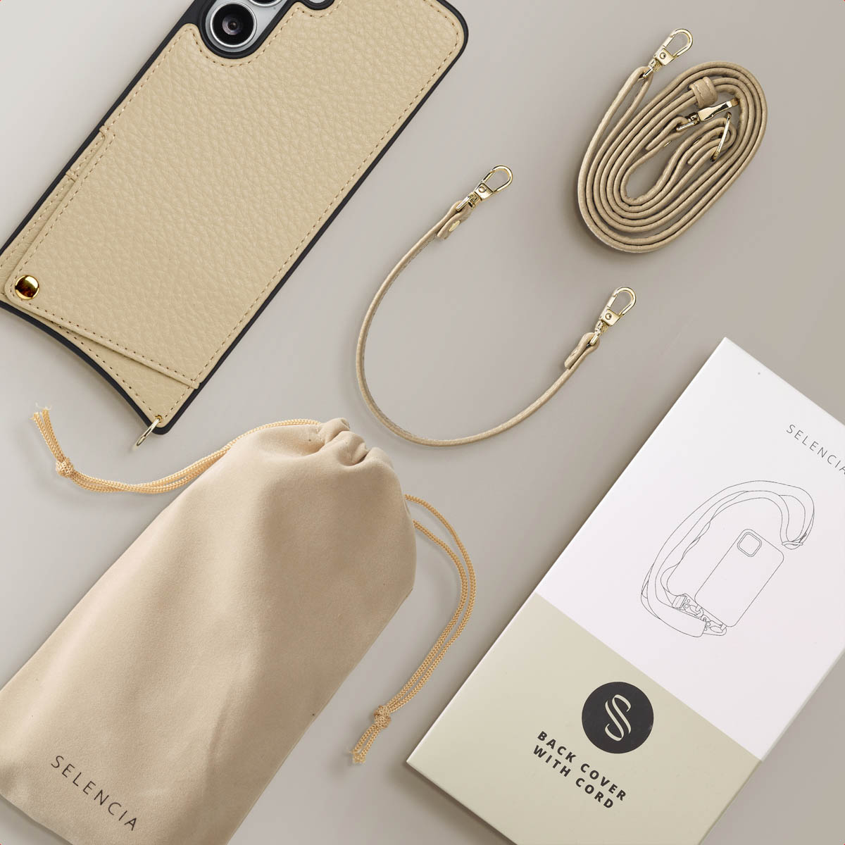 Een beige telefoonhoesje, koord, zakje en verpakking zijn gerangschikt op een lichtgrijze ondergrond. De telefoonhoes heeft een camera en een zwarte rand.