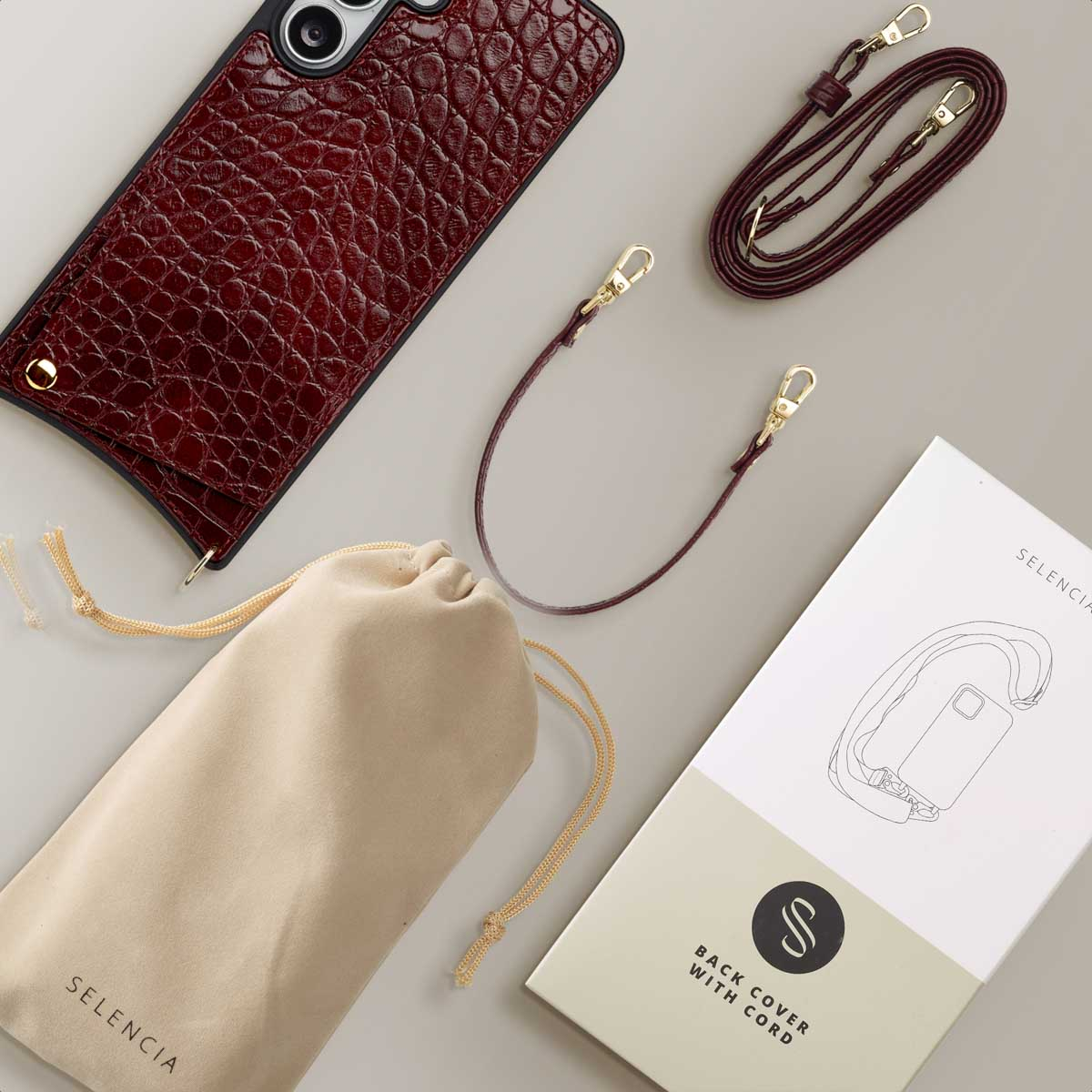 Een set telefoonaccessoires met hoes, tas, riem en handleiding op een grijze ondergrond. De hoes is bordeaux.