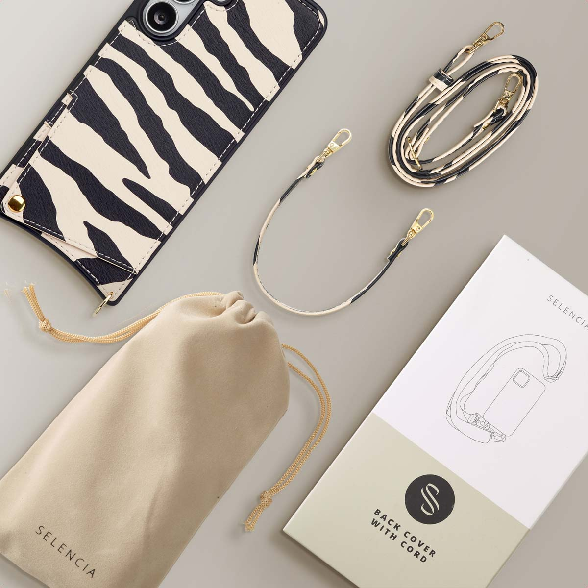 Telefoonhoesje met zebra strepen, een tas, een riem en een handleiding liggen op een grijze ondergrond. Alle items hebben het merk SELENCIA.