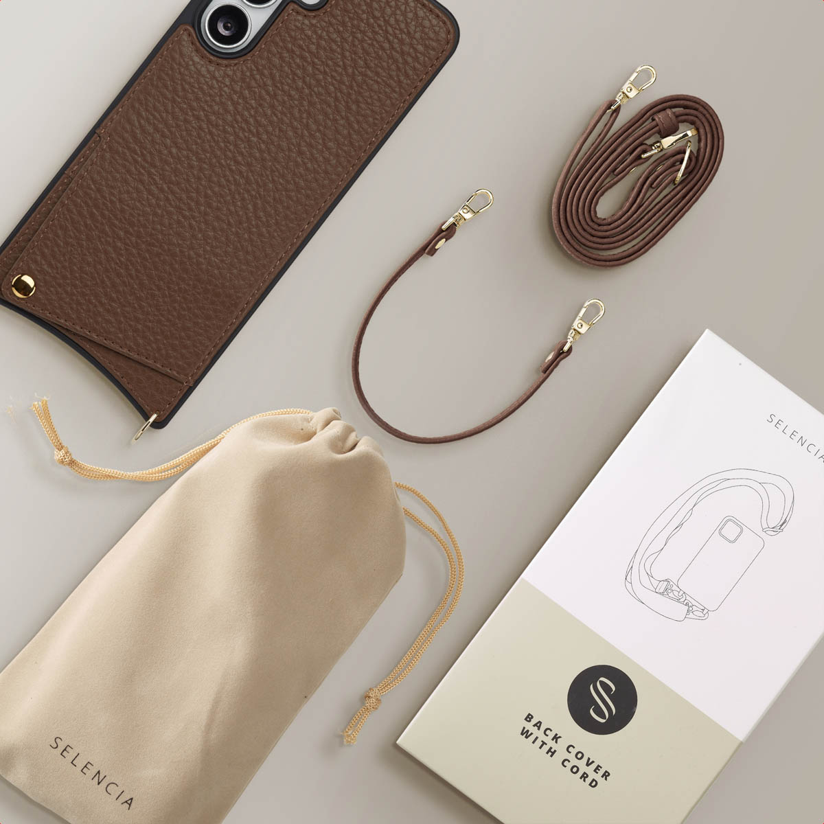 Een bruine telefoonhoes, riemen, een tas en een doos, allemaal op een beige oppervlak. Op de doos staat 'Back Cover with Cord'.