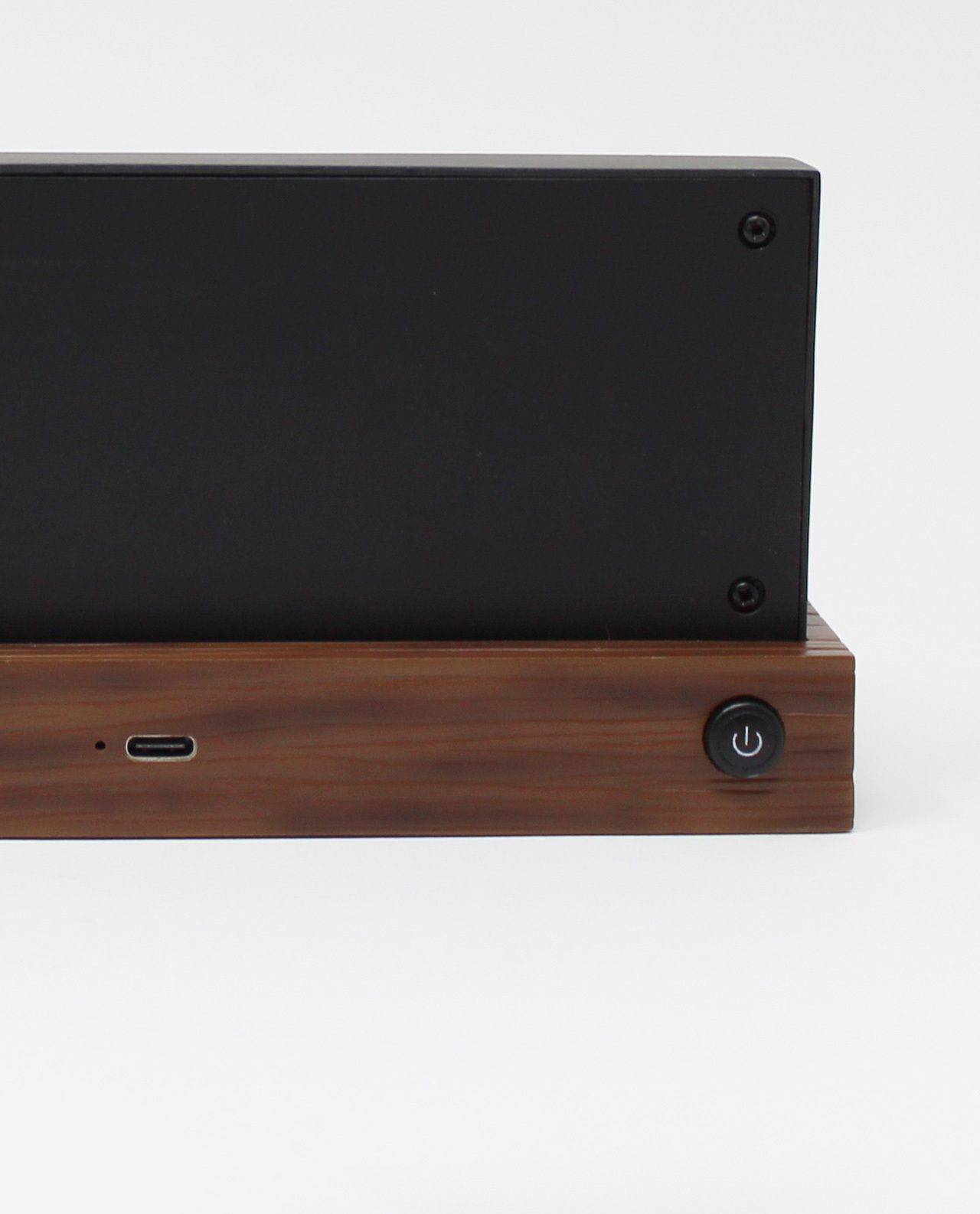 Un dispositivo nero su una base di legno. Pulsante di accensione e porta USB-C visibili. Sfondo bianco.