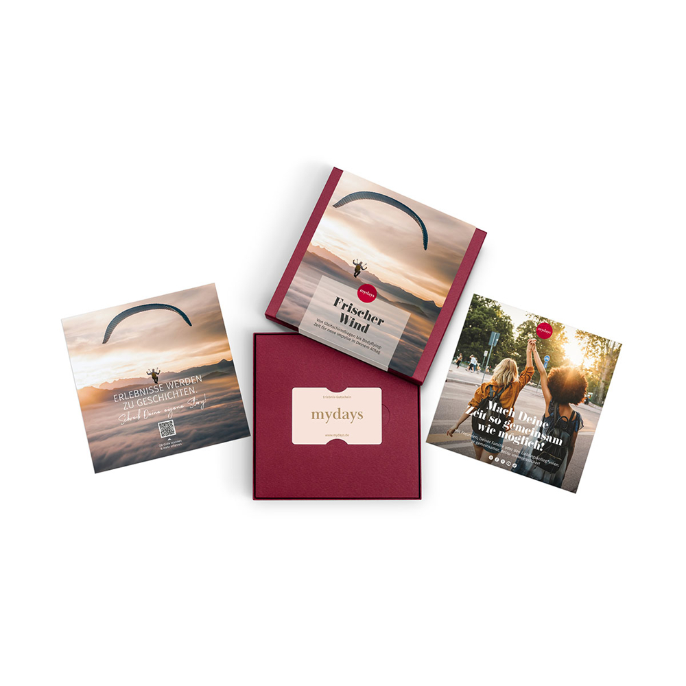 Vier mydays-Geschenkboxen und -Karten sind auf einer weißen Oberfläche verteilt. Sie zeigen Bilder von Gleitschirmen und Frauen mit deutschem Text.