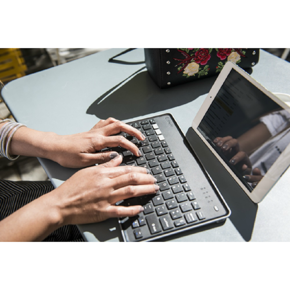 Mani che digitano su una tastiera nera. Sono visibili anche un tablet, una borsa con motivo floreale e un tavolo.