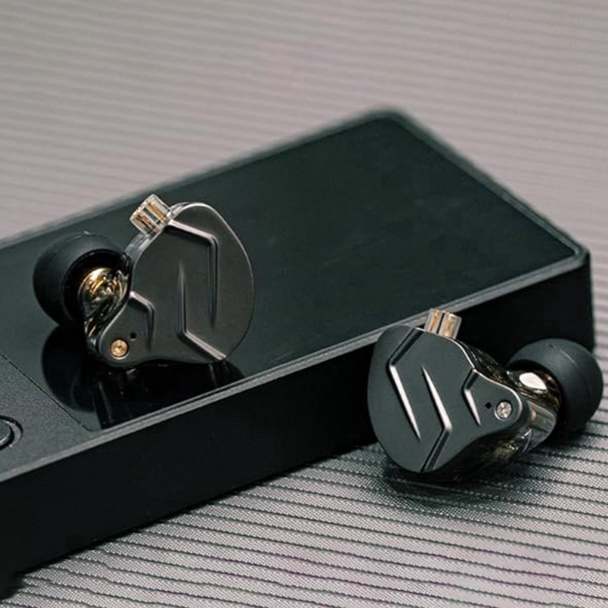 Twee zwarte en zilveren in-ear-hoofdtelefoons op een zwart apparaat, op een grijs oppervlak met strepen. De hoofdtelefoons hebben gouden accenten.