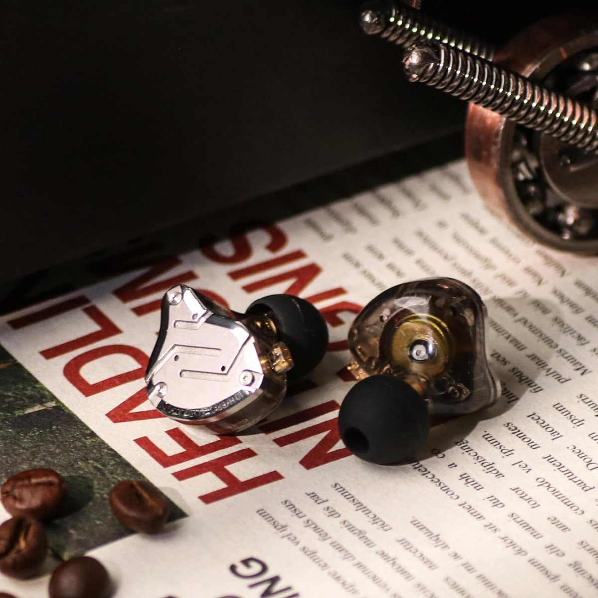 Twee in-ear koptelefoons op een krant met koffiebonen. Eén is zilver, de andere is transparant met zwarte uiteinden.