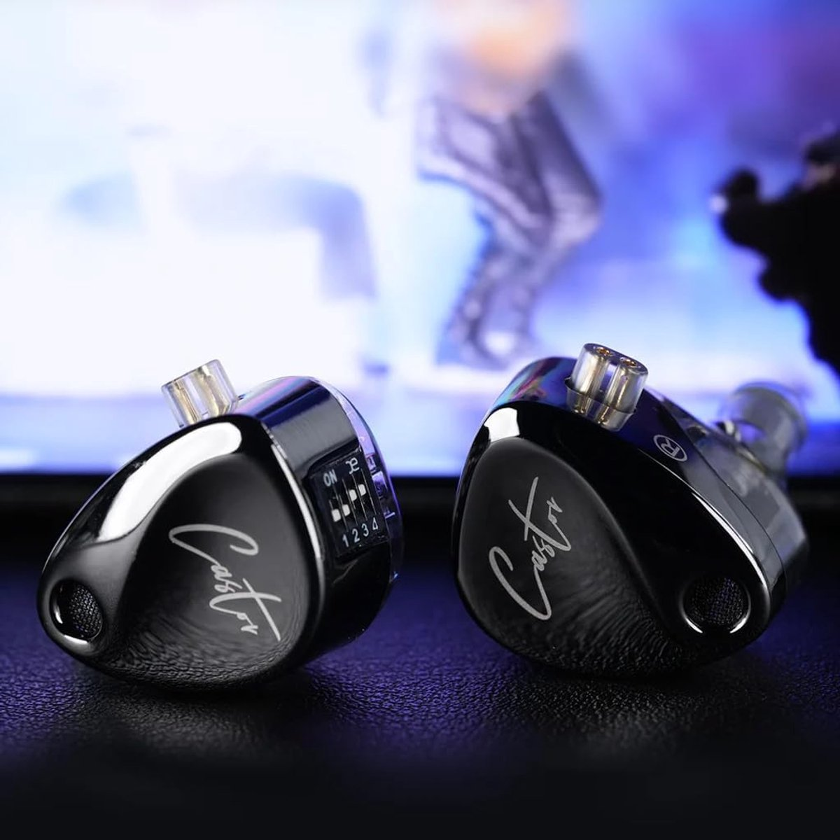 Two black in-ear headphones with the name 'Castor' on them. A switch with numbers is visible. The background is blurred.