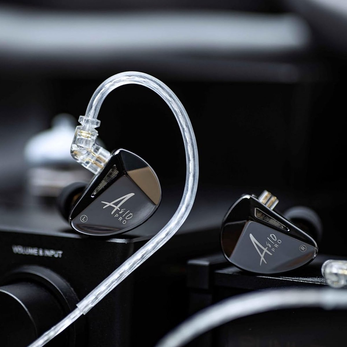 Two black and silver in-ear monitors with clear cables. One is in focus, the other is blurred. Dark background.