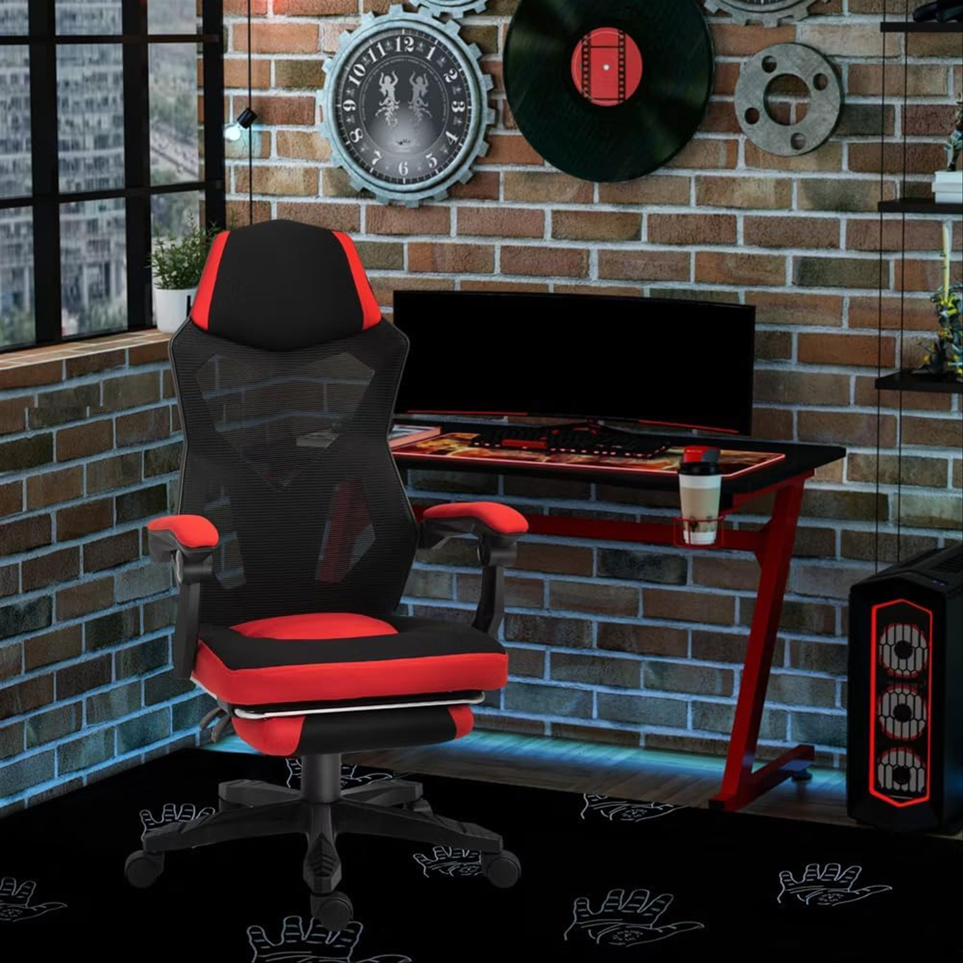 Una configuración de juego con una silla roja y negra, una computadora, un escritorio y un fondo de pared de ladrillos.