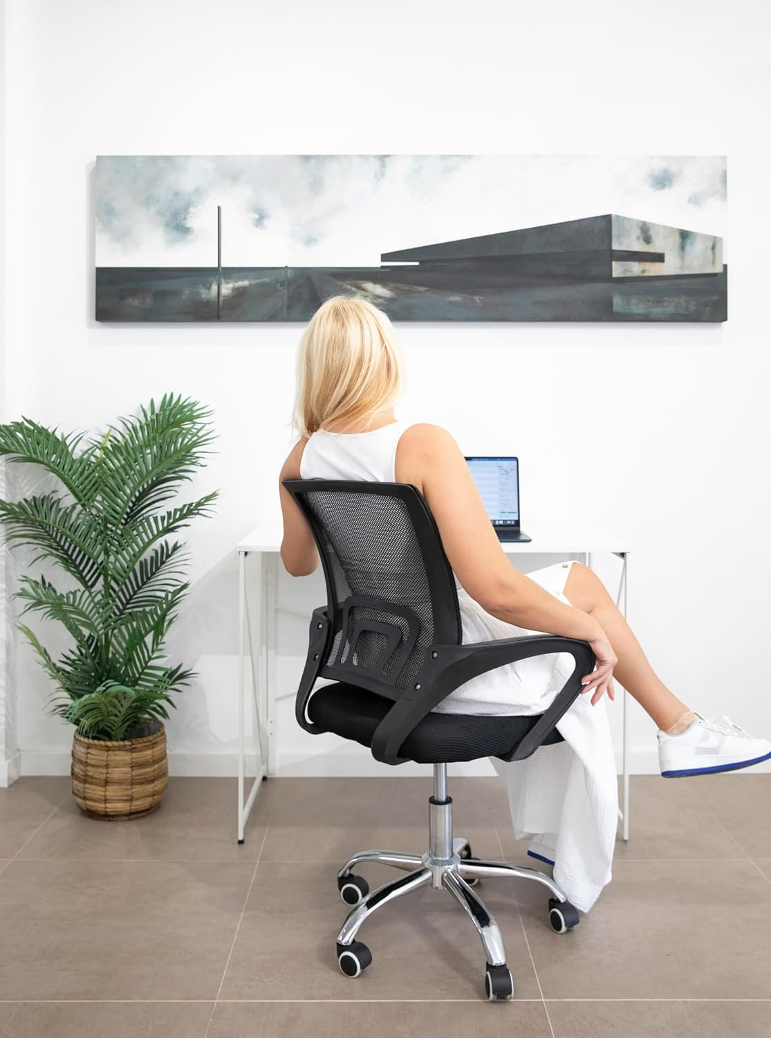 Mujer de vestido blanco sentada en escritorio con portátil, de espaldas. Planta en maceta a la izquierda. Pintura en la pared blanca.