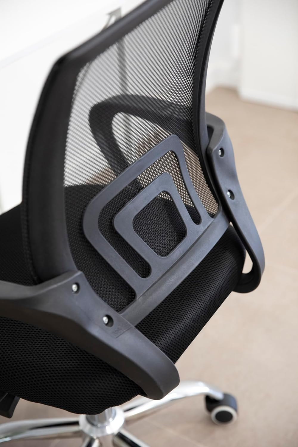 Primer plano de una silla de oficina negra. Tiene respaldo de malla, marco de plástico y base cromada con ruedas sobre suelo claro.