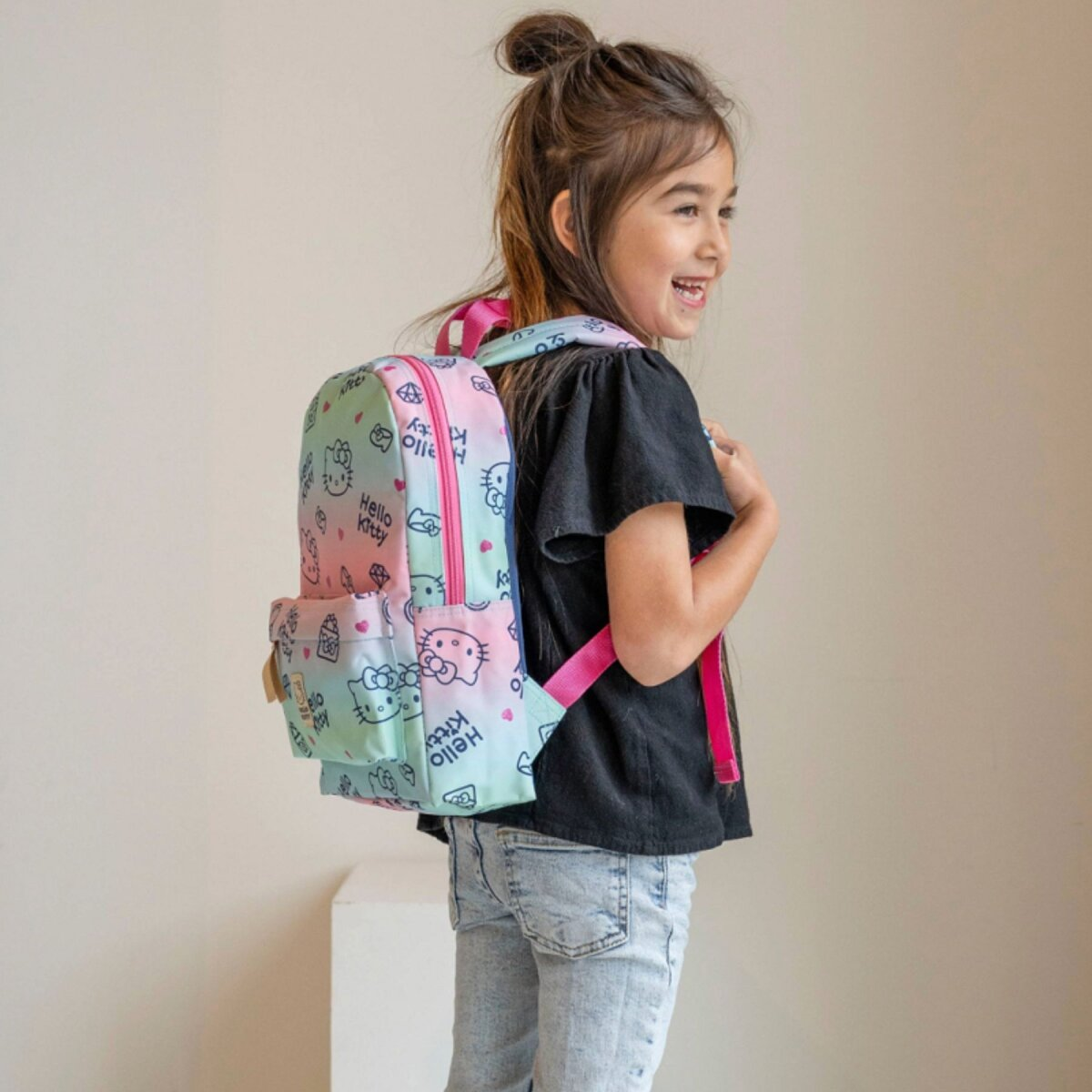 A girl with a Hello Kitty backpack smiles. She wears a black top and jeans. The background is a plain, light wall.