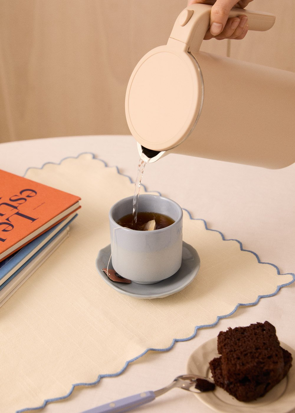 Una persona vierte agua de una tetera beige en una taza de té azul. Un pastel y un libro están cerca. El mantel tiene ribete azul.