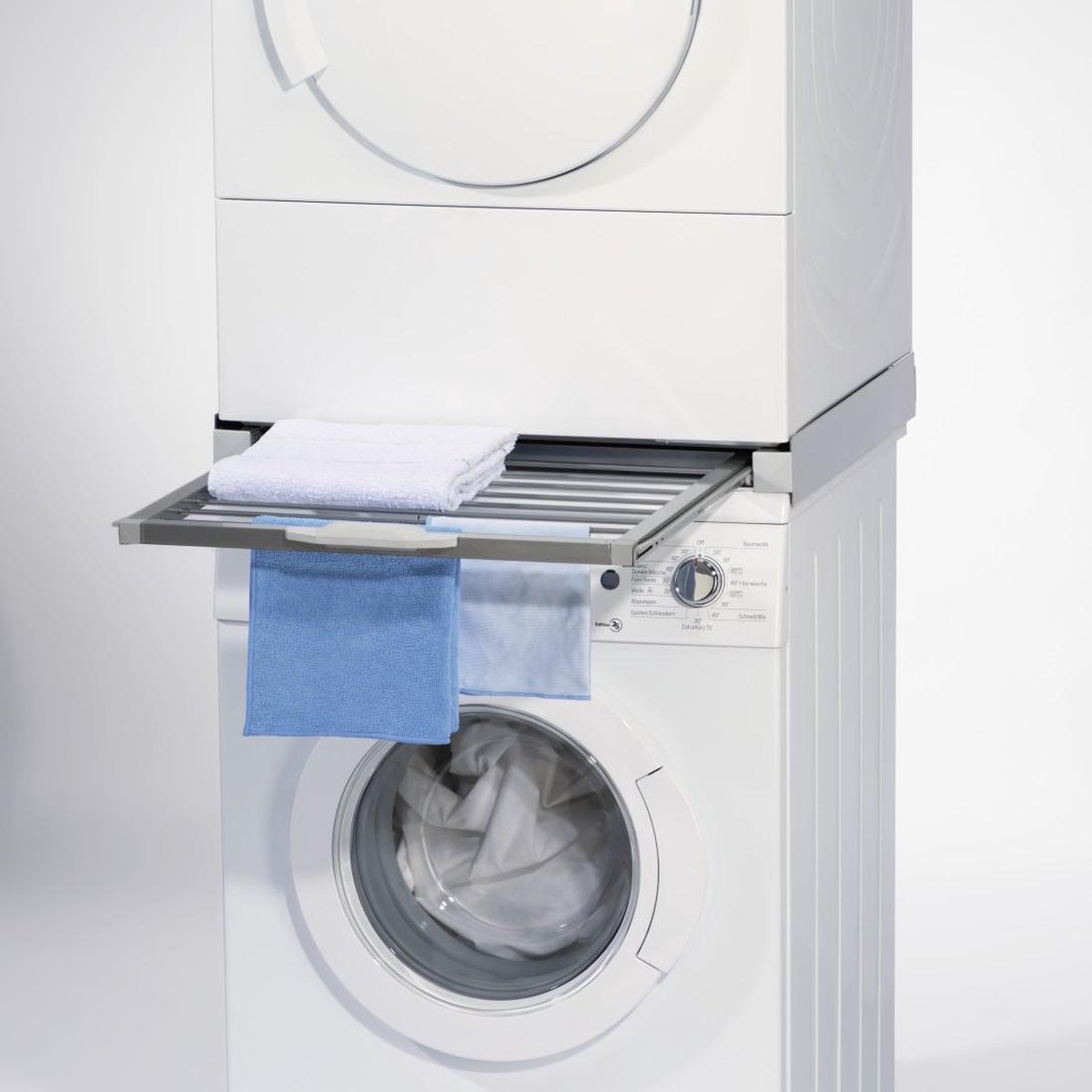 Eine weiße Waschmaschine mit einem Trockner oben. Ein Gestell hält ein weißes Handtuch und zwei blaue Handtücher. Die Waschmaschine ist an.
