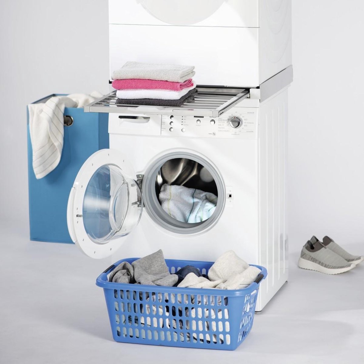 Eine Waschmaschine mit offener Tür, ein blauer Korb mit Kleidung, Handtüchern und eine blaue Wäschesack. Sneakers sind rechts.