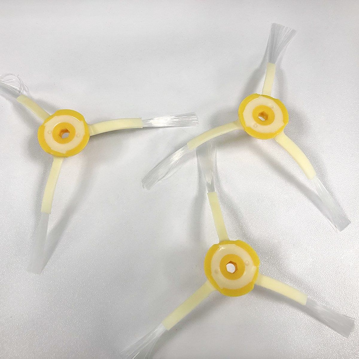 Trzy żółto-białe szczotki do robota sprzątającego leżą na białej powierzchni. Każda ma trzy ramiona i centralne koło.