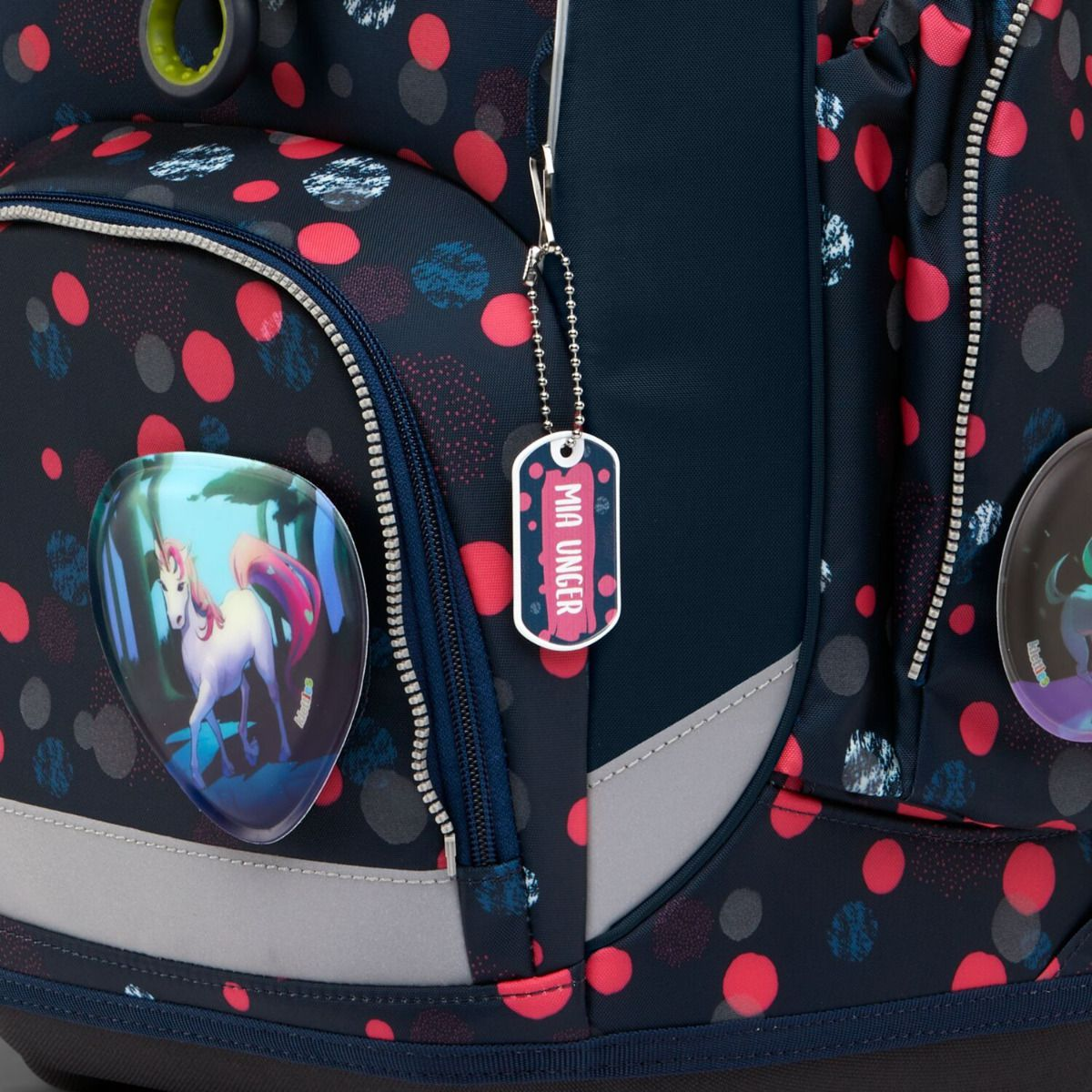 Ein marineblauer Rucksack mit roten Punkten hat ein Einhorn-Emblem. Ein Anhänger mit 'Mia Unger' hängt daran. Details sind Reißverschlüsse und reflektierende Streifen.